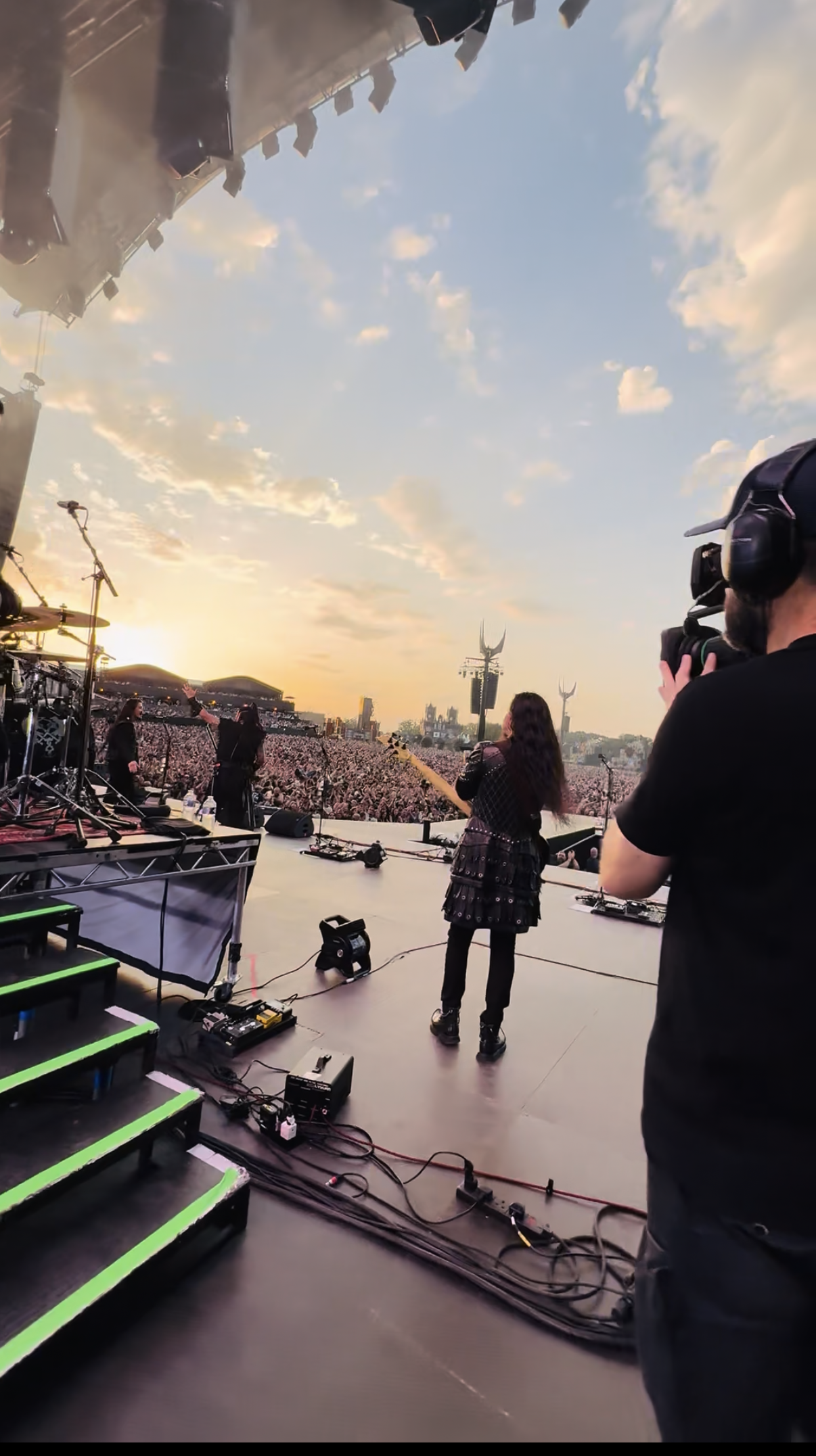 Concert stage at sunset with performers and a large crowd in the background. A camera operator is filming, and sound equipment is visible on stage.