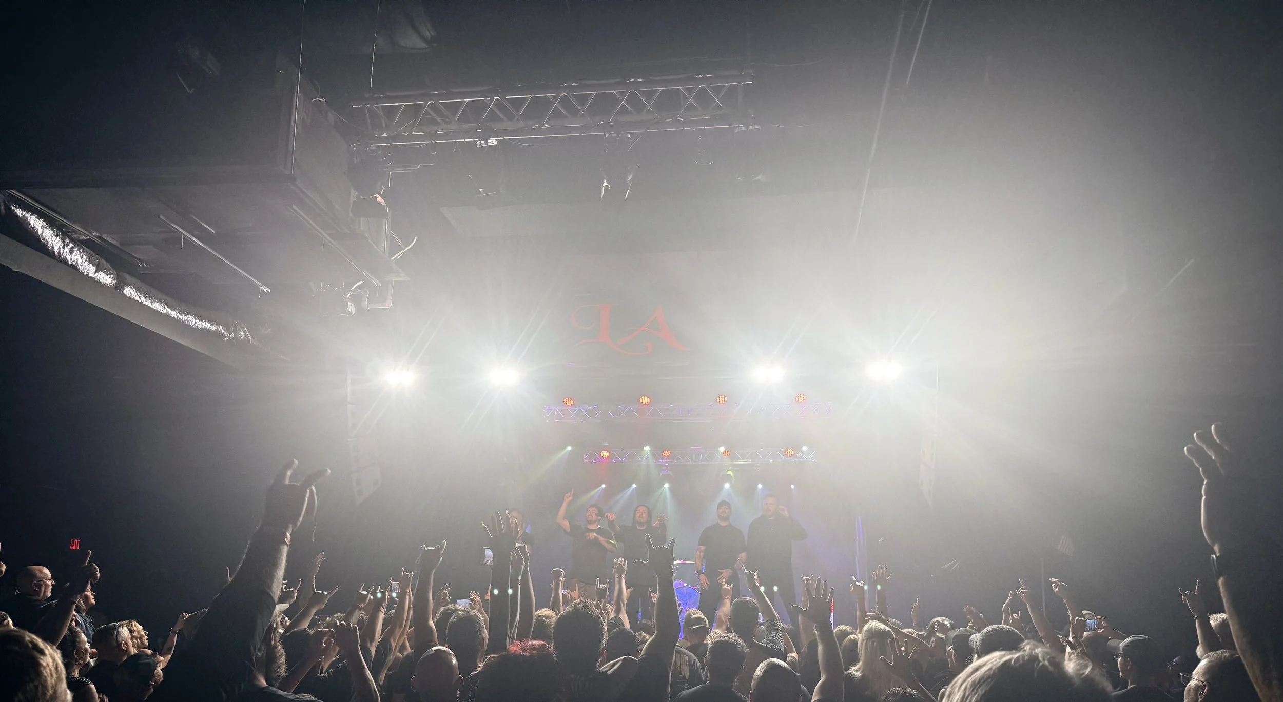 Concert scene with performers on stage and audience raising hands, bright stage lights, and a large screen with red initials. AR Pro Audio is running FOH.