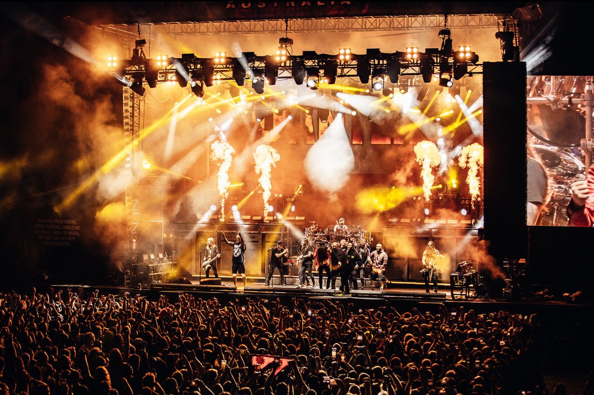 A live concert scene with a band performing on stage, surrounded by bright yellow and white lights, smoke effects, and pyrotechnics, with a large crowd of fans in the foreground.