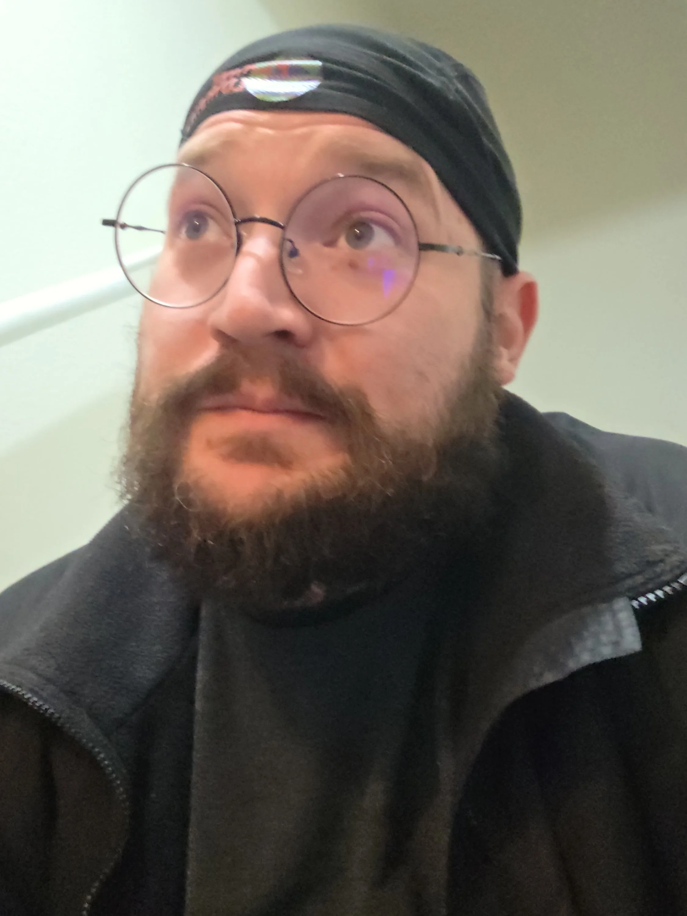 Close-up of a man with a beard wearing round glasses, a black beanie, and a black jacket, looking upward.