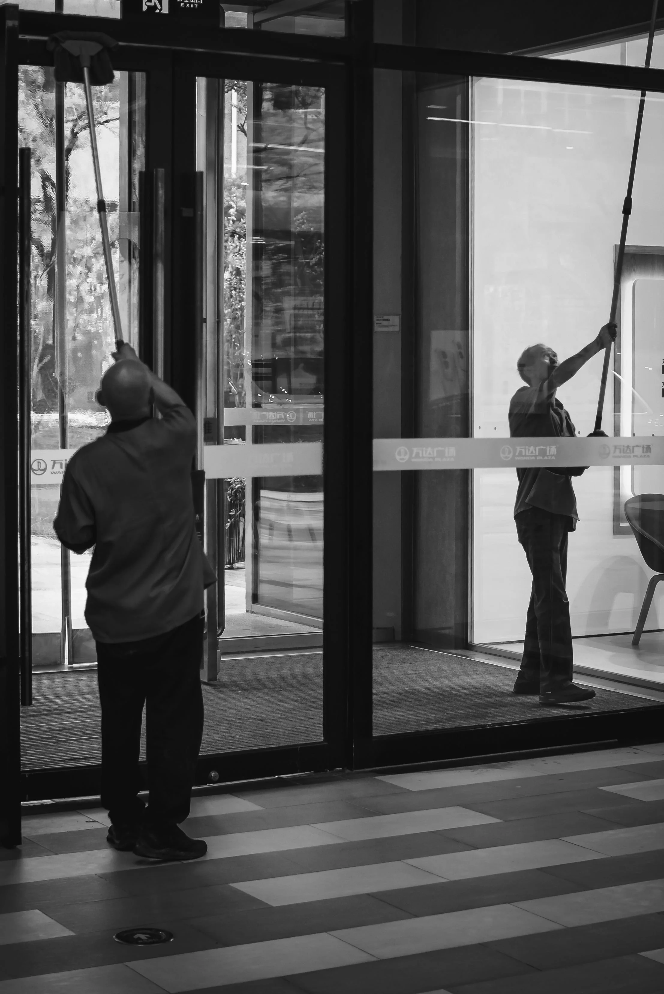 Black and white photo of three people cleaning glass doors in a shopping mall. Two are inside, one is outside, using scrapers with long handles. The mall has glass doors, sliding open, with visible signage. The floor has a segmented tile pattern.