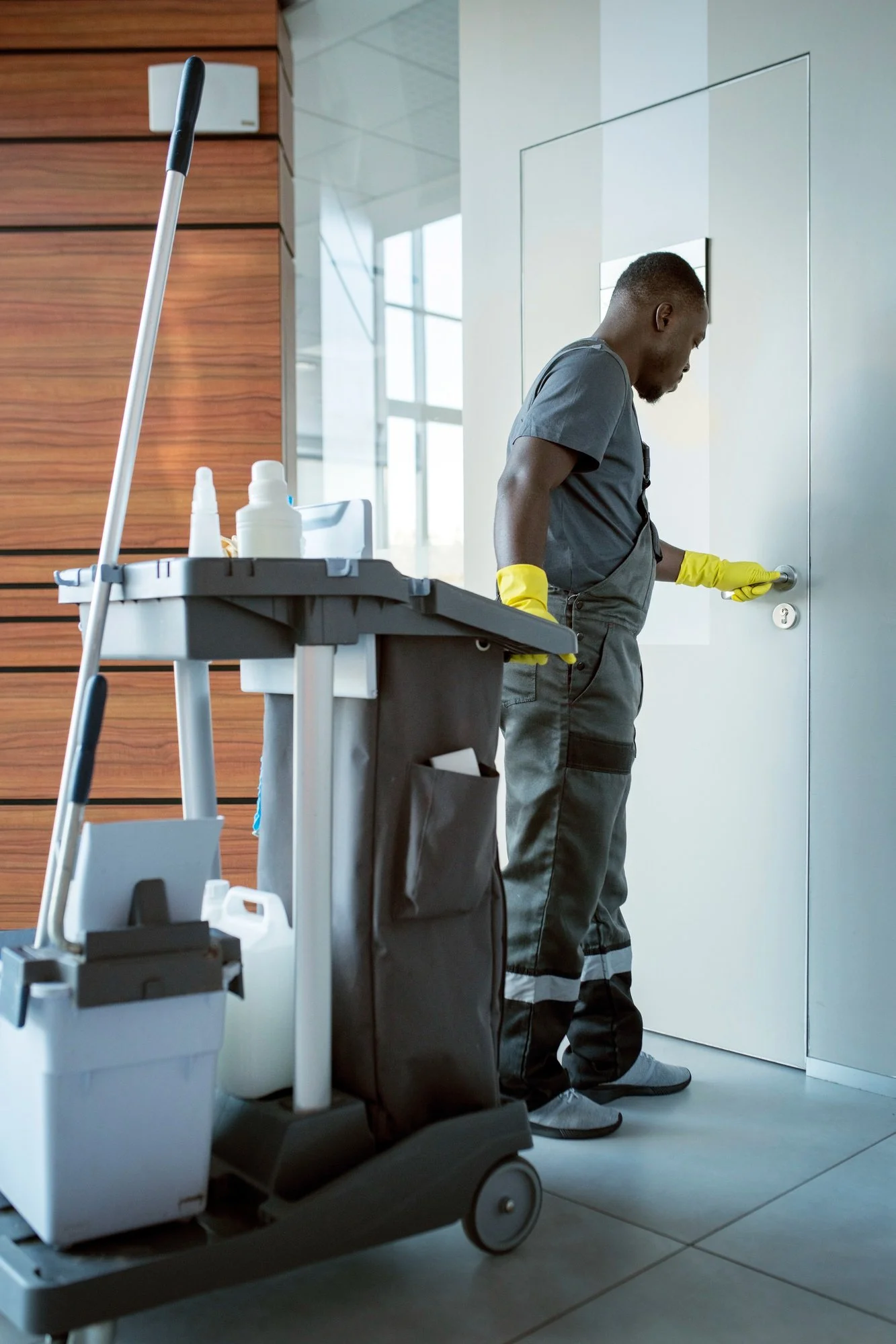 Janitor wearing yellow gloves operating a door handle in a modern building lobby, with cleaning supplies on a cart nearby.
