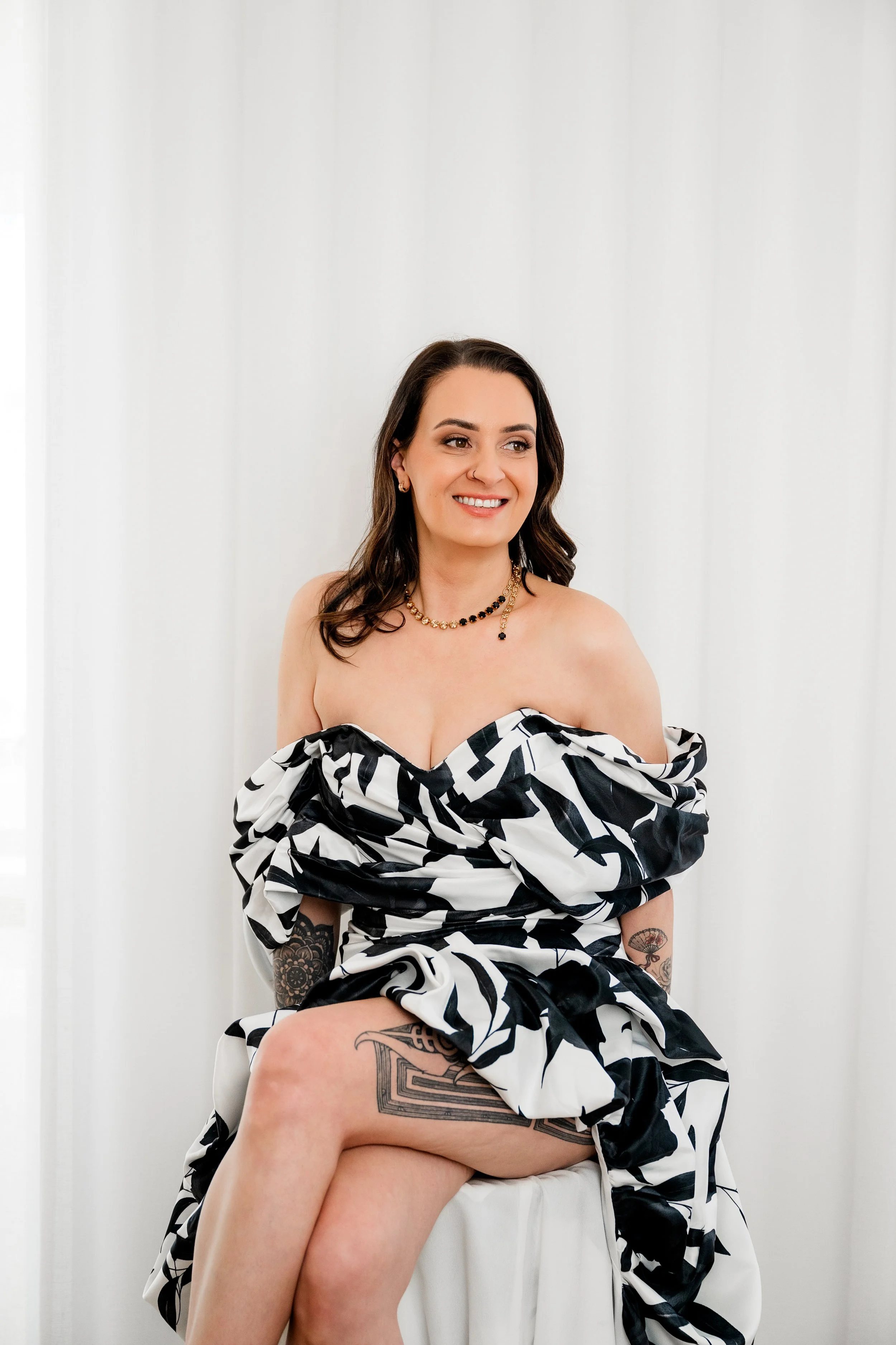A woman sitting on a white chair against a plain white wall, smiling and looking to the side. She is wearing a black and white patterned dress, gold jewelry, and has visible tattoos on her arms and legs.