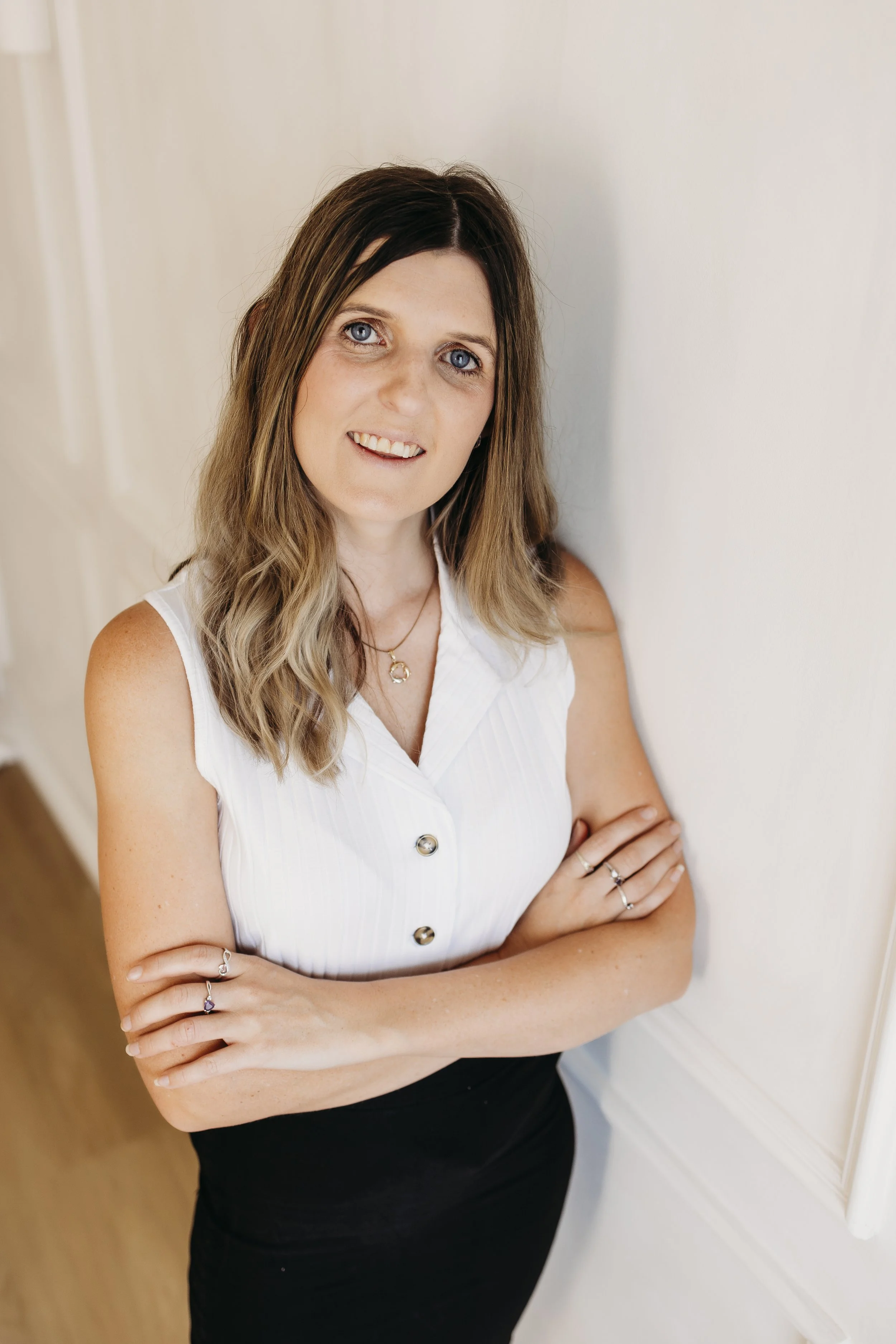 A woman with shoulder-length brown hair and blue eyes, wearing a sleeveless white shirt with buttons, standing with arms crossed, smiling softly, against a white wall.
