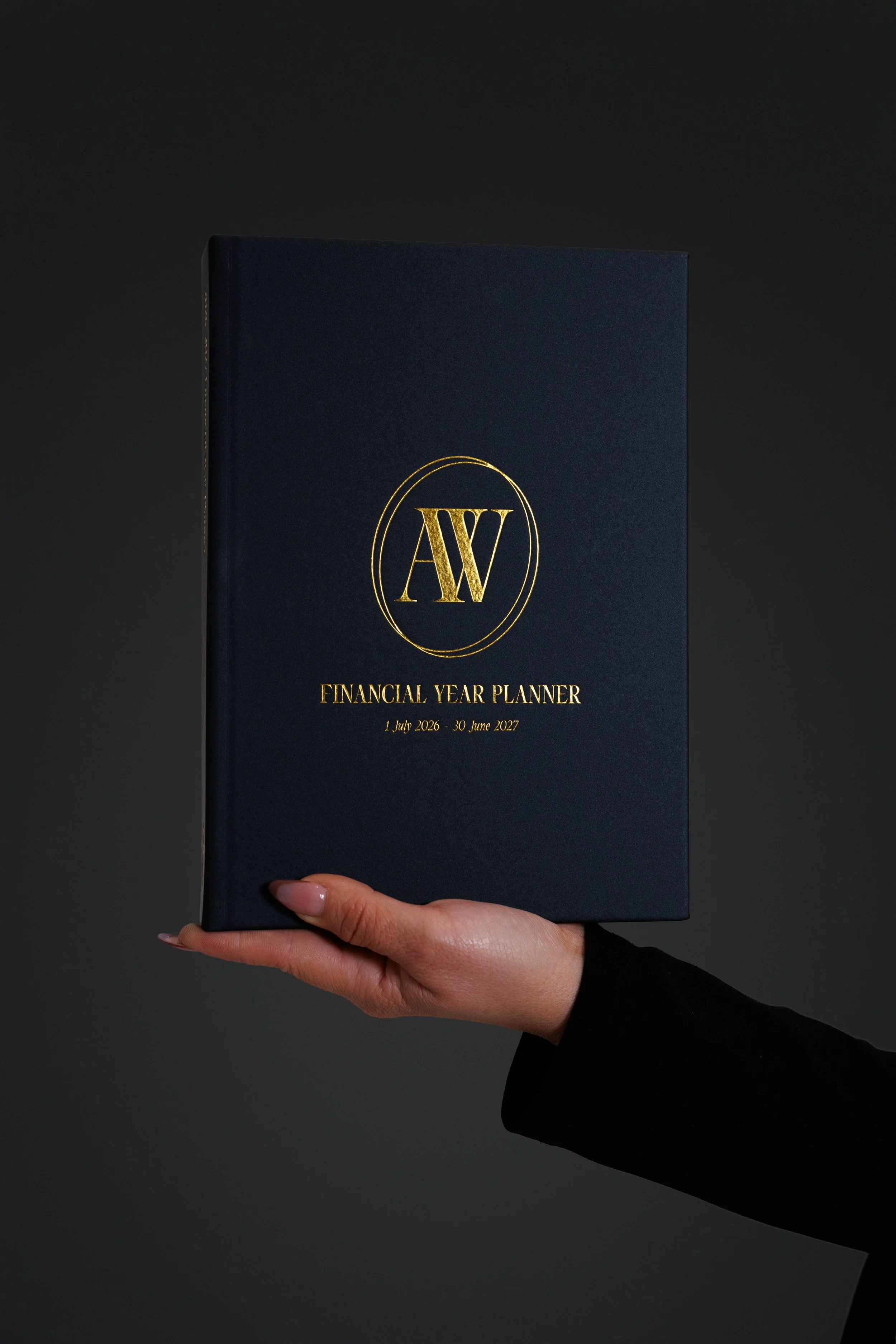 A person with a black long sleeve shirt is holding a navy blue book titled 'Financial Year Planner' with gold lettering on a dark background.