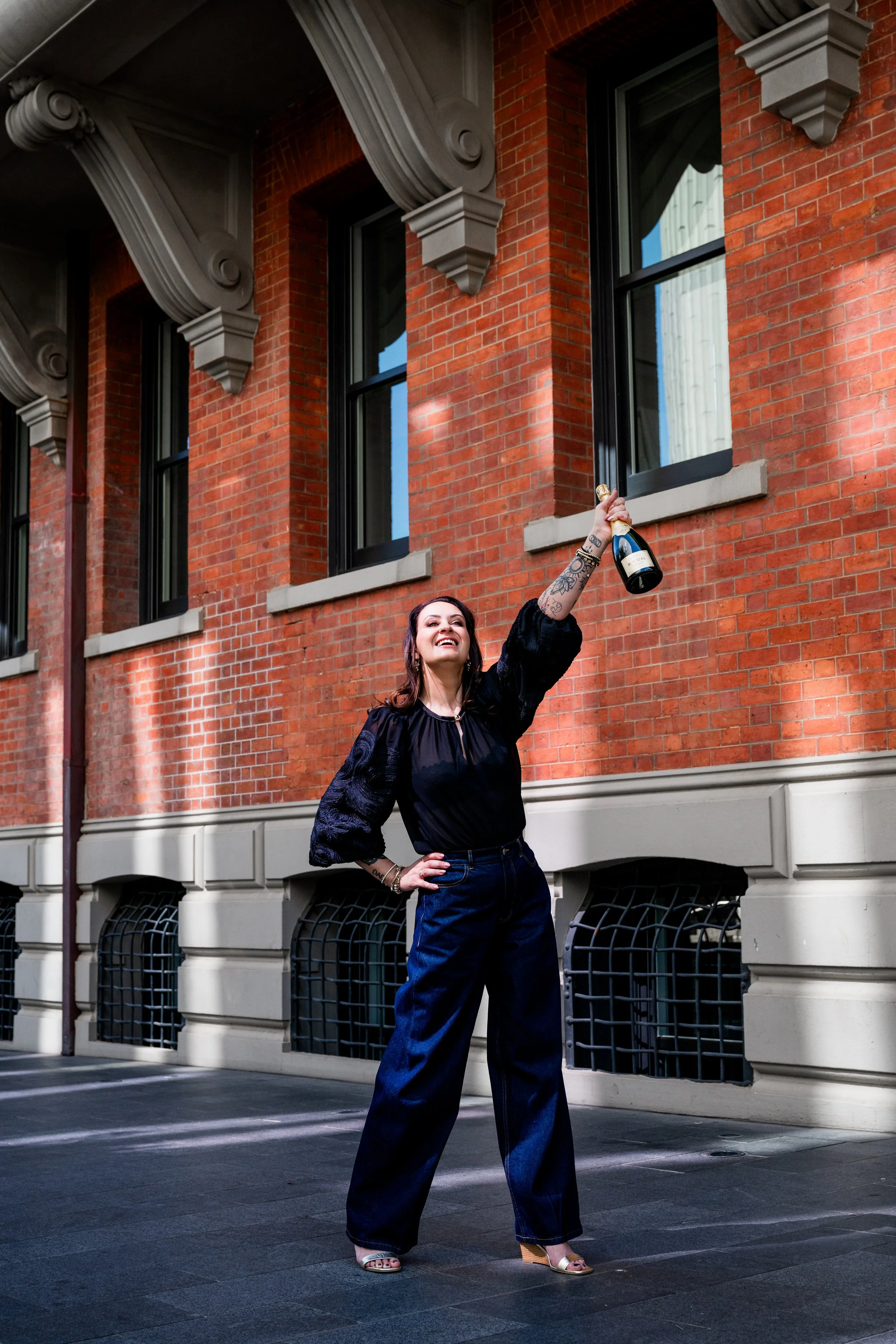 A woman with tattoos and long hair, wearing a black top and wide-leg jeans, stands on a city street holding a bottle of champagne overhead in celebration, in front of a brick building with large windows.