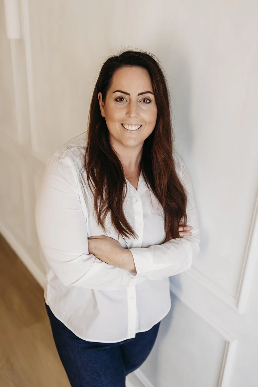 A woman with long brown hair, wearing a white button-up shirt and jeans, standing with arms crossed and smiling at the camera in a brightly lit room.