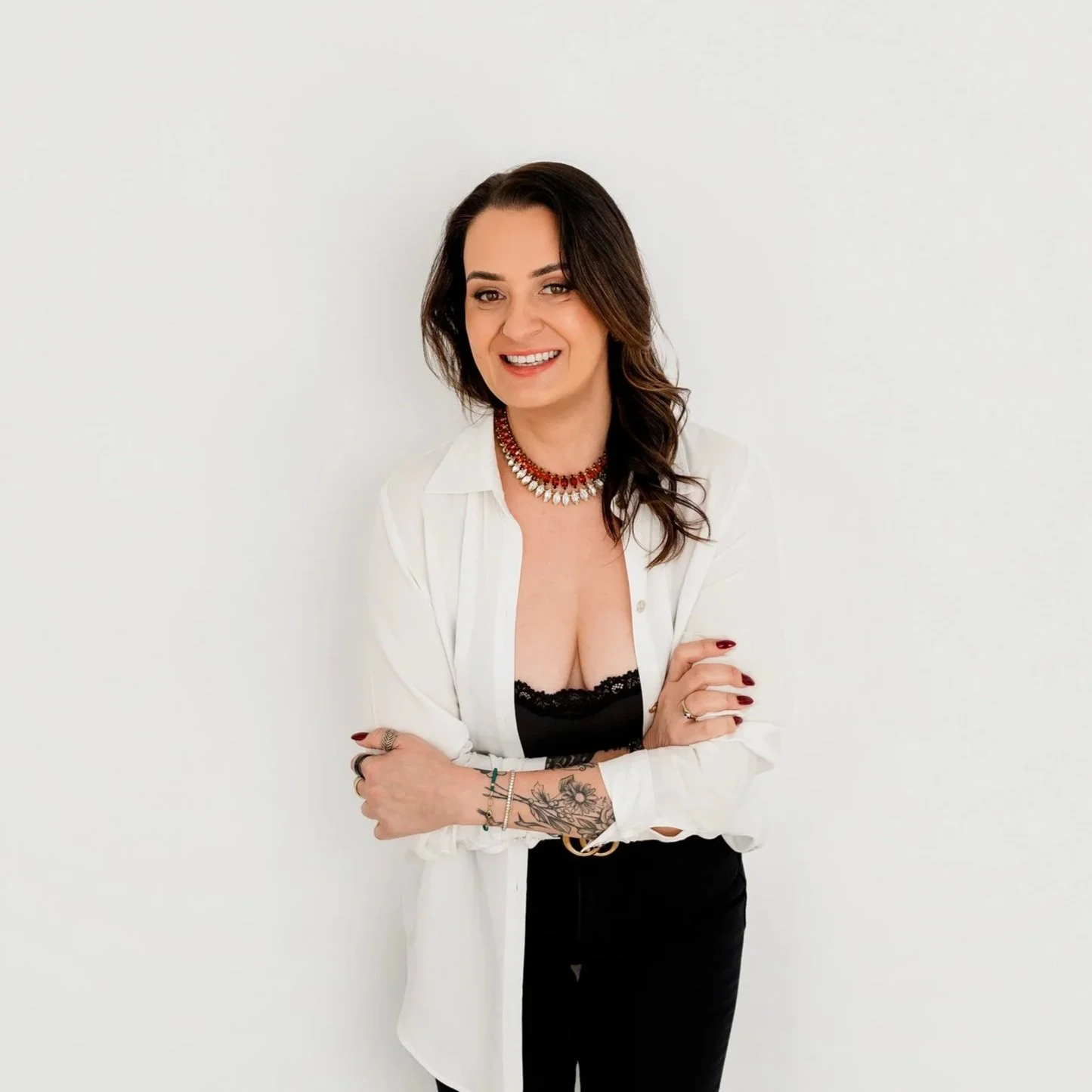 Woman with tattoos and dark brown hair, smiling, wearing a white shirt over black lace top, red beaded necklace, standing against white wall.