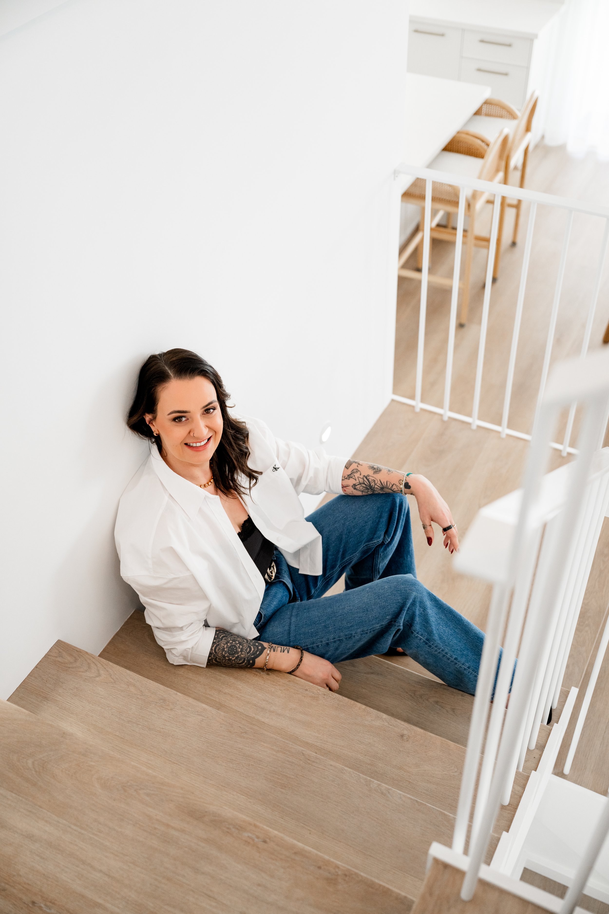 A woman with dark hair, wearing a white shirt and blue jeans, sitting on wooden stairs in a bright room, smiling at the camera.