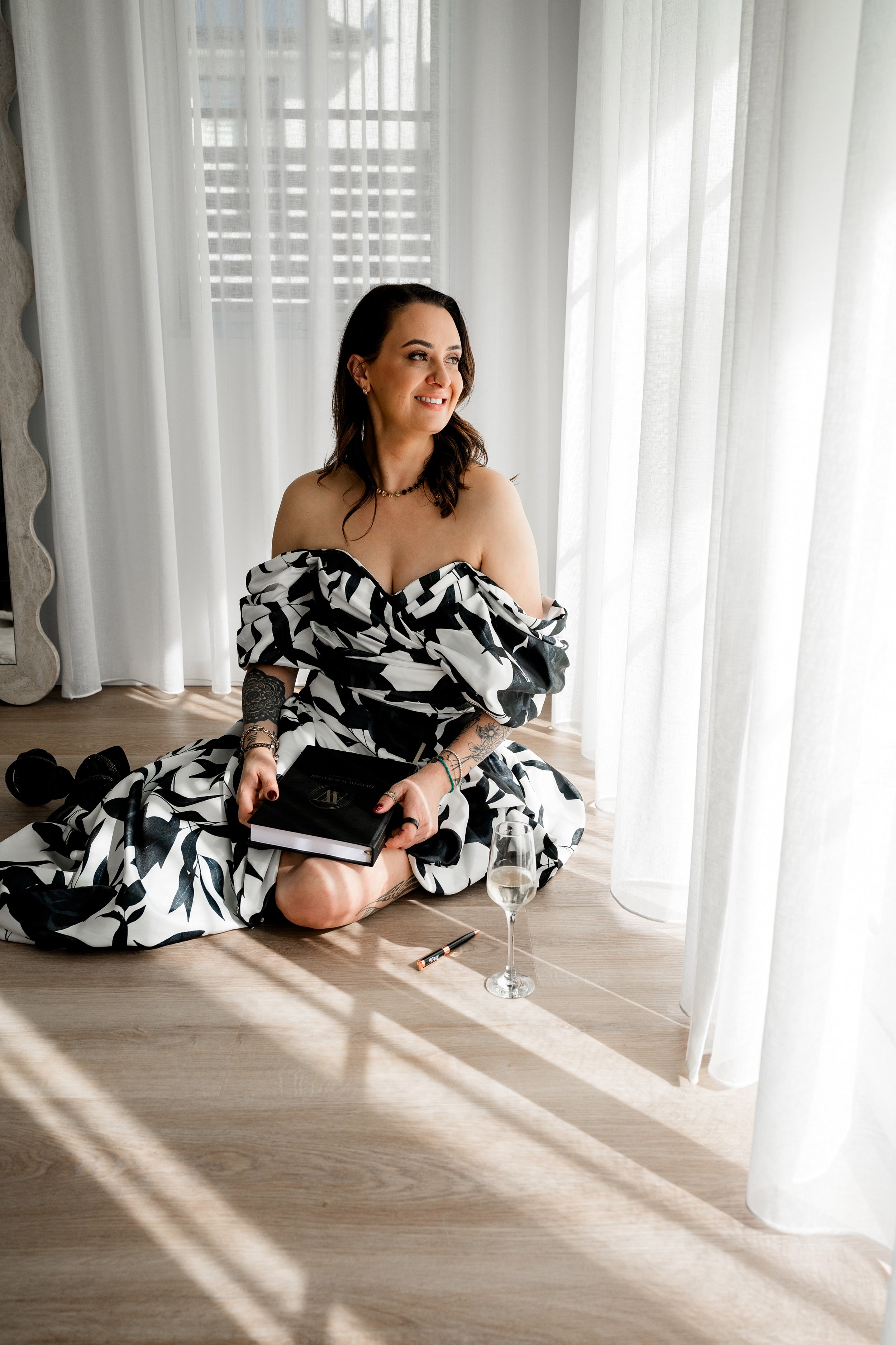 A woman sitting on the floor next to a window with white curtains, smiling and holding a book. She has tattoos on her arm and is dressed in a black and white patterned off-shoulder dress. There is a glass of champagne, a lipstick, and a pen on the floor nearby.