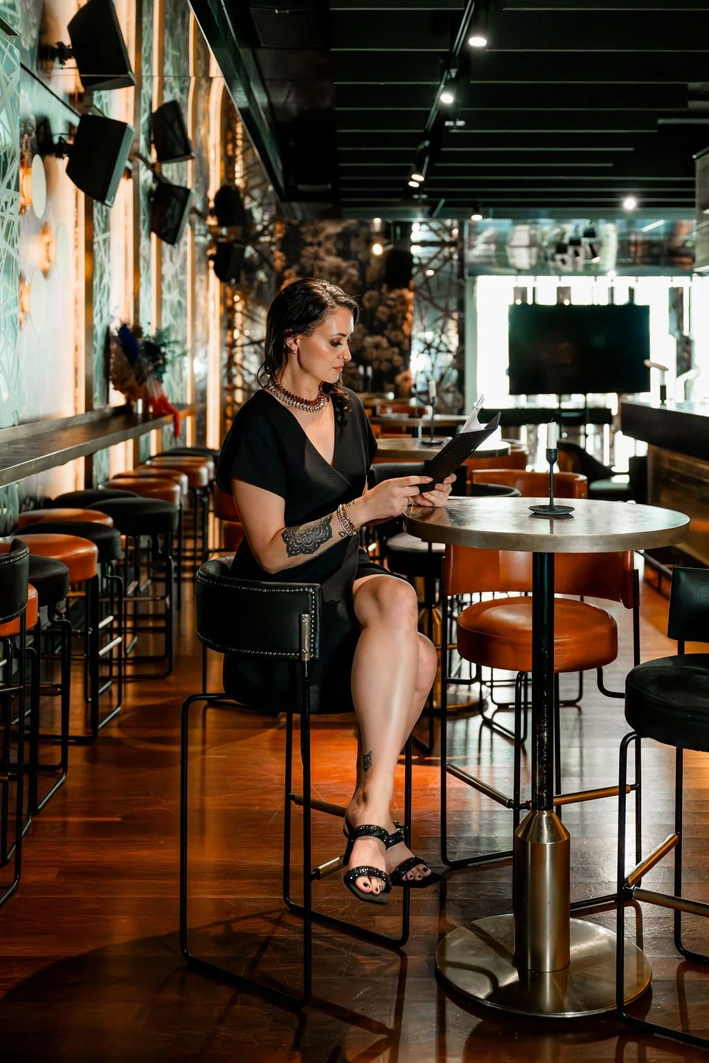 A woman sitting alone at a high table in a dimly lit restaurant or bar, reading a menu or tablet, surrounded by bar stools, with a large TV screen and decorative wall art in the background.