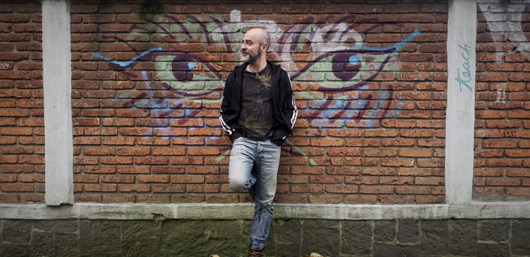 G.Kowalsky Author in a black jacket and jeans is standing in front of a brick wall with colorful graffiti of a large pair of eyes. He is smiling and looking off to the side.