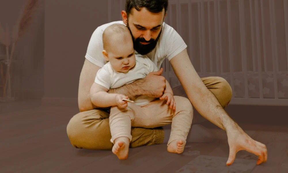 A man and a baby sitting on the floor, with the man holding the baby, who is holding a wooden stick. The background shows a room with vertical blinds.