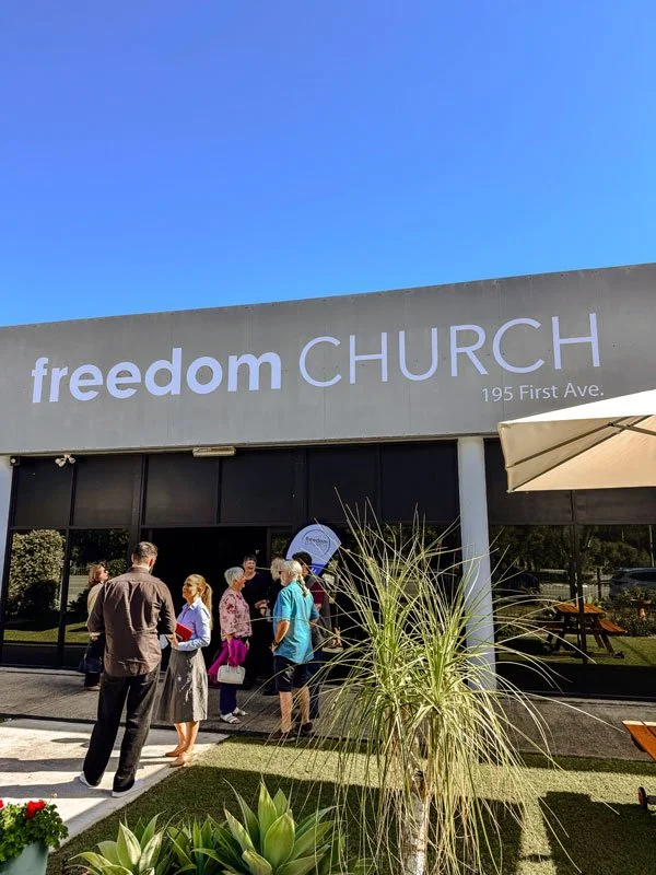 People standing outside the entrance of Freedom Church at 195 First Ave, with a clear blue sky above and plants in the foreground.