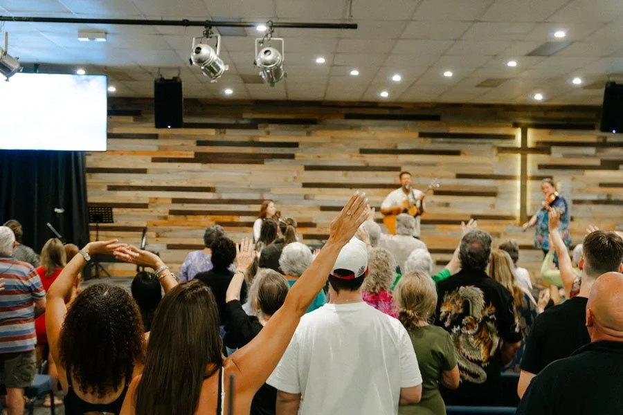 People gathered in a church or community center, raising hands during a musical performance with two singers and a guitarist on stage.