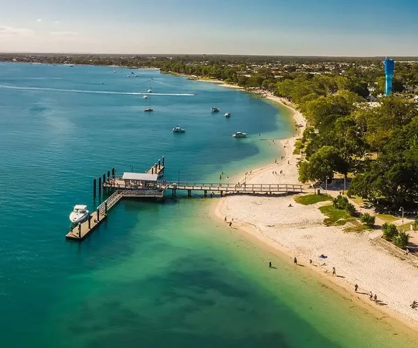 Aerial view of a beach with a pier extending into a calm, turquoise water. Several boats are anchored near the shore, surrounded by green trees and a cityscape in the background.