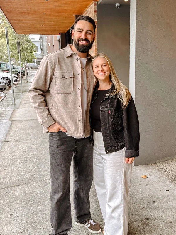 A man and woman standing on a sidewalk outside a building, smiling at the camera. The man has a beard and is wearing a beige jacket over a light shirt and dark jeans. The woman has long blonde hair, wearing a black jacket over a black shirt and white pants. There are parked cars and trees in the background.