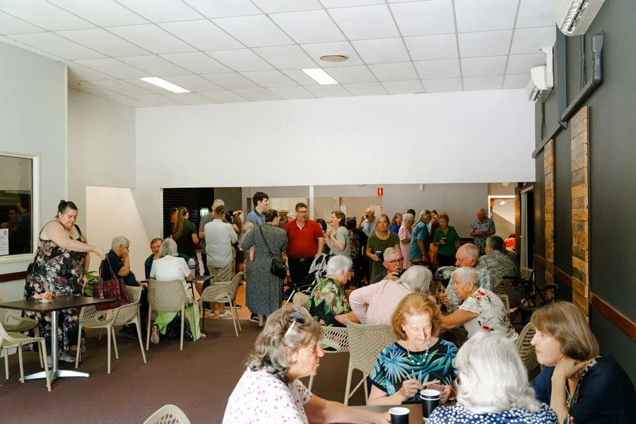 A gathering of mostly older adults in a community hall, some sitting and some standing, engaging in conversations and activities.