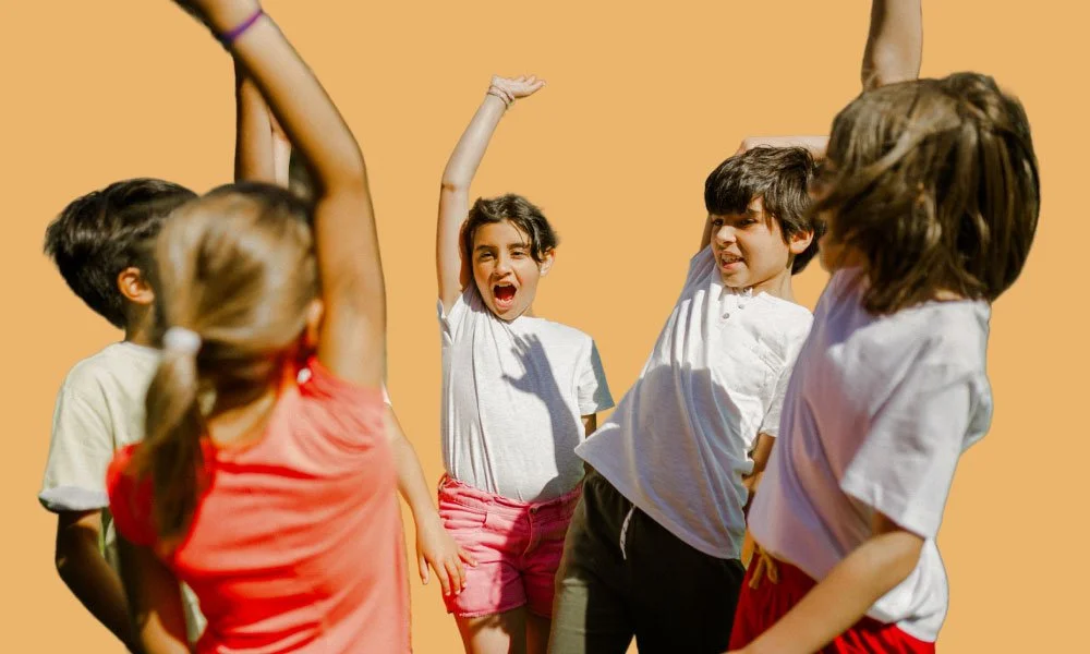 A group of children are playing and laughing outside on a sunny day, some with one hand raised.
