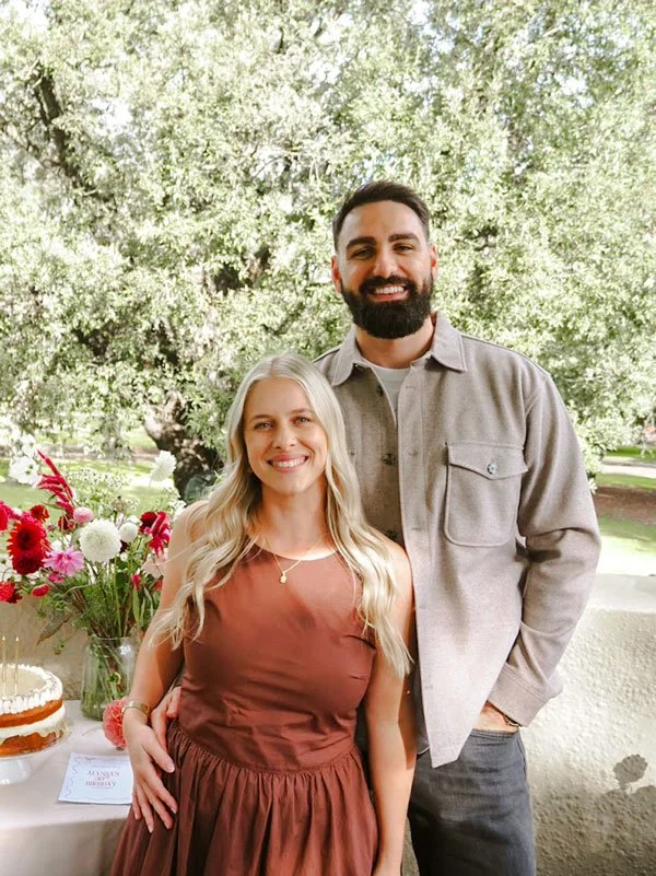 A smiling man and woman standing outdoors near a table with a cake and flowers, with trees and grass in the background.