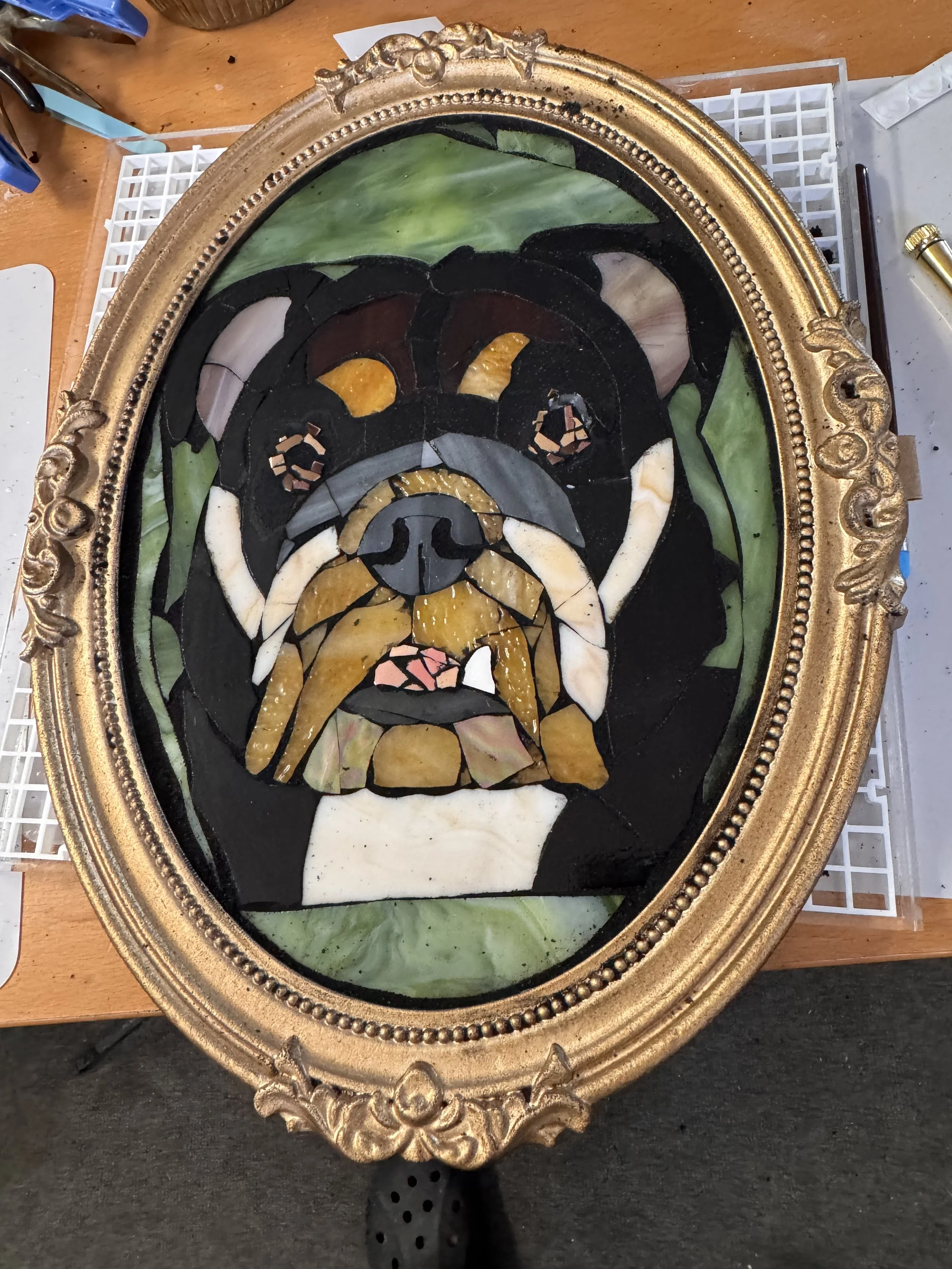 An ornate, oval-shaped picture frame containing a colorful stained glass mosaic of a dog's face, placed on a workbench surrounded by tools and supplies.