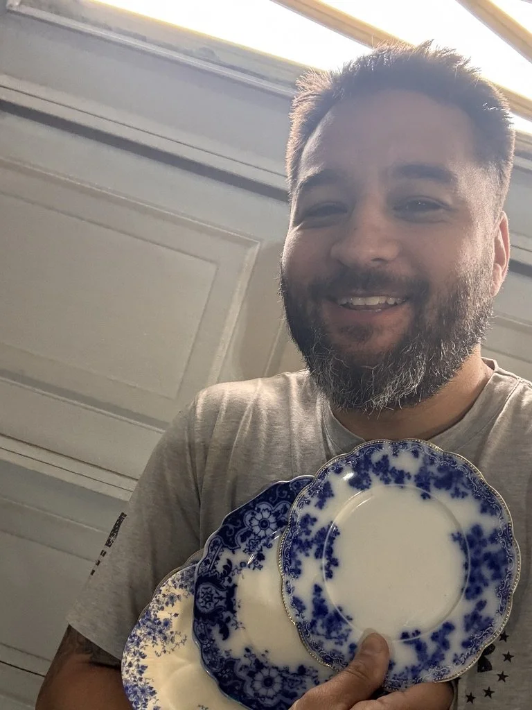 A man with a beard smiling while holding several blue and white decorative plates in front of him. The plates have floral and intricate patterns. The background shows a white door and a window.