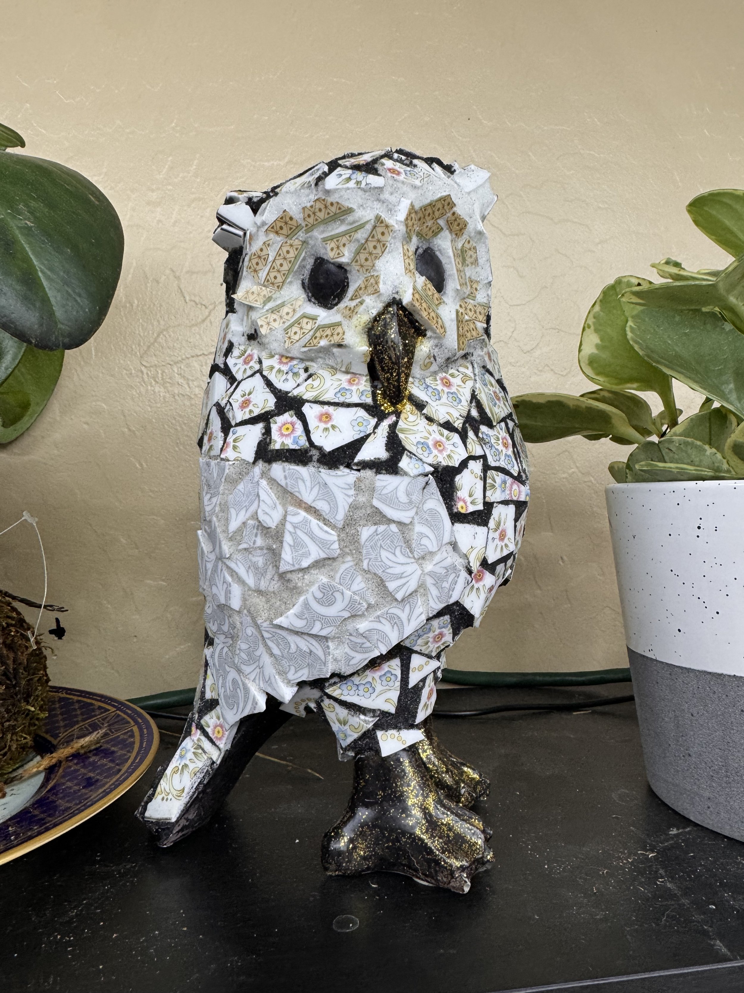 A mosaic sculpture of a dog made from broken porcelain pieces, with black eyes and a gold-tipped nose, sitting on a black surface beside a potted plant.
