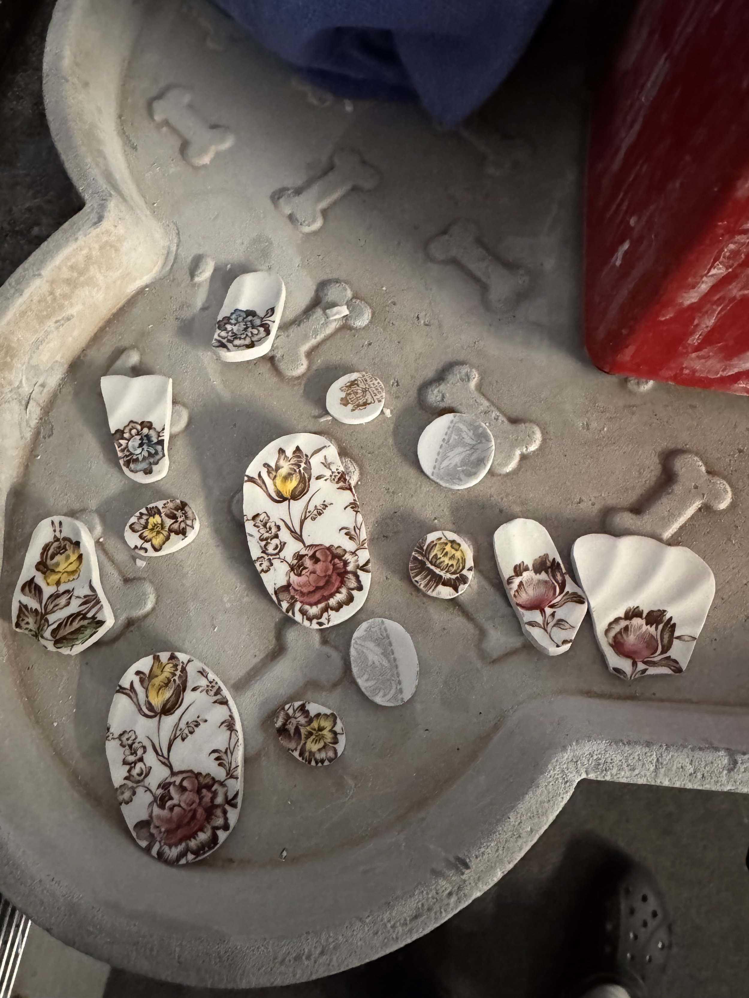 Broken ceramic pieces with floral patterns on a white surface, scattered on a stone or concrete surface. Part of a red object and a gray footwear are visible.