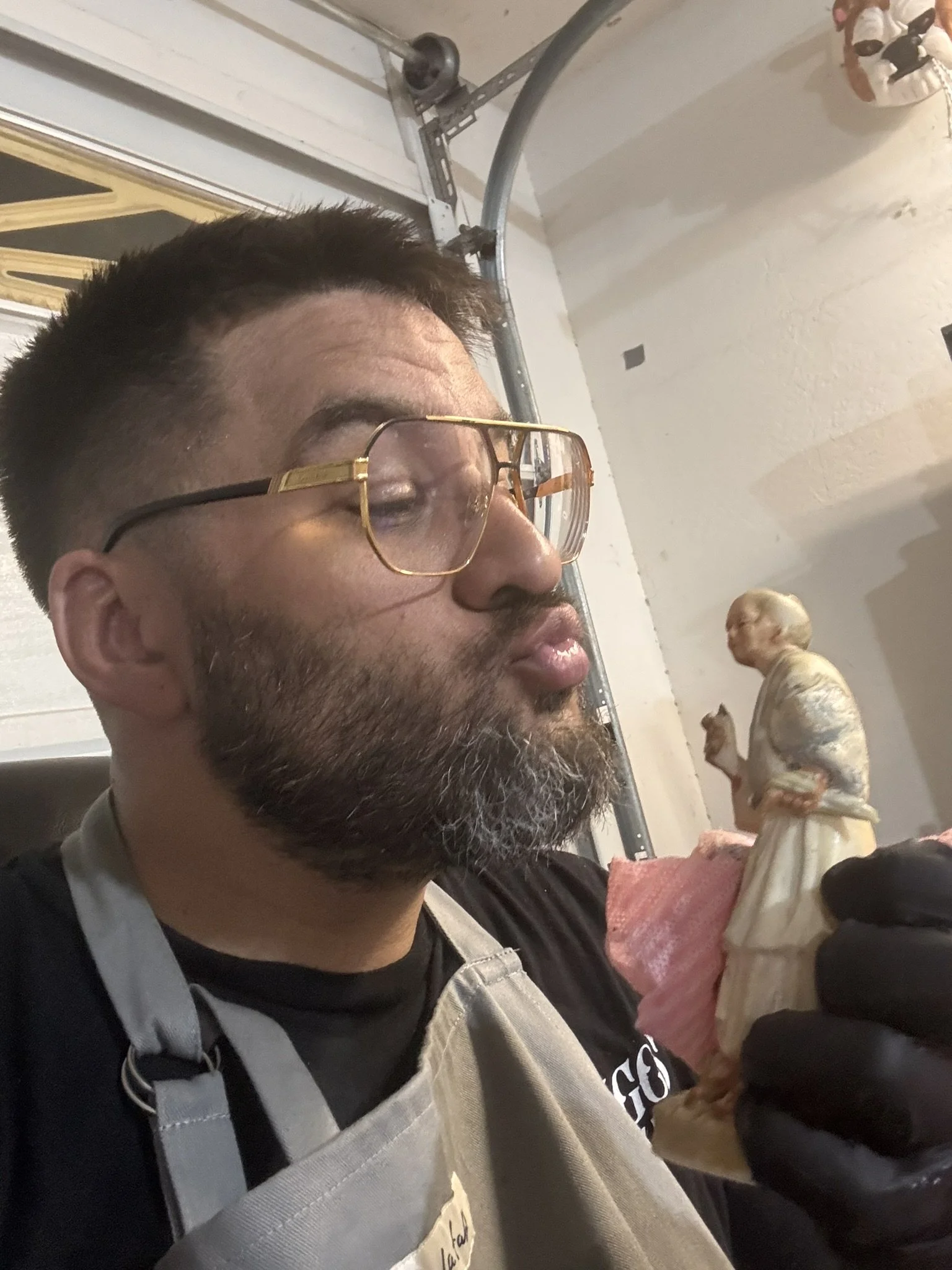 A close-up of a man with glasses and a beard, wearing an apron and a black shirt, sitting with sliced raw meat and a butcher's knife in front of him. In the background, there is a person in a white apron holding a piece of meat.