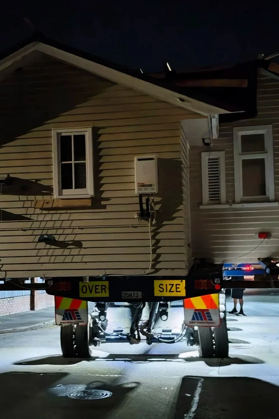A weatherboard home mounted on an oversize transport trailer navigating a residential street late at night during a house removal project.