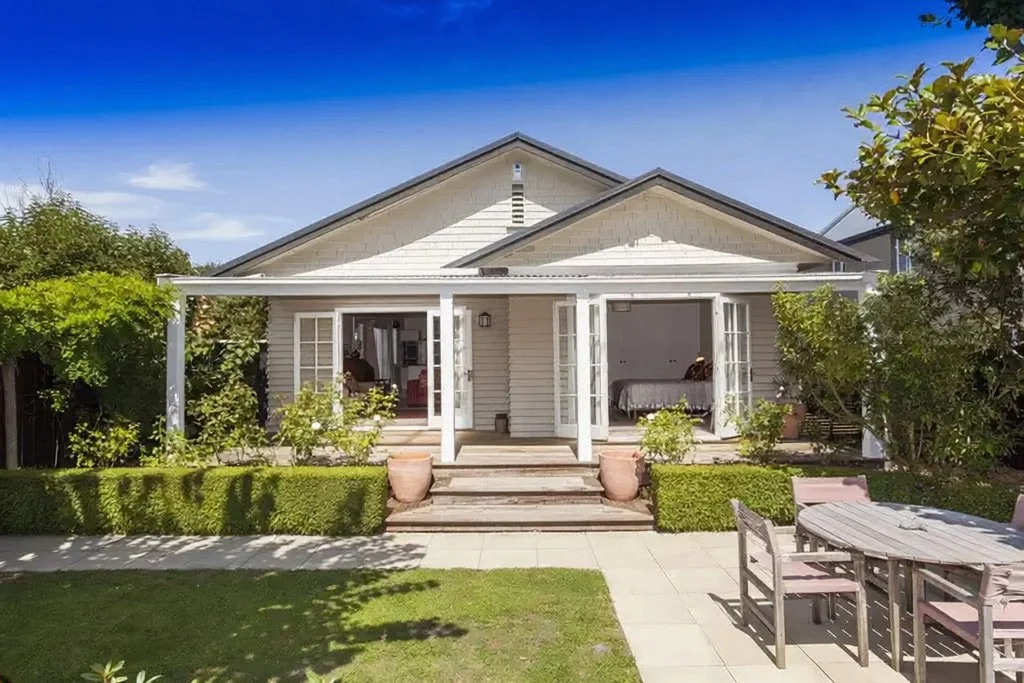 Christchurch house removal project showing the exterior of a charming, refurbished two-story timber bungalow with French doors, a manicured garden with a patio and dining set, under a bright blue sky.
