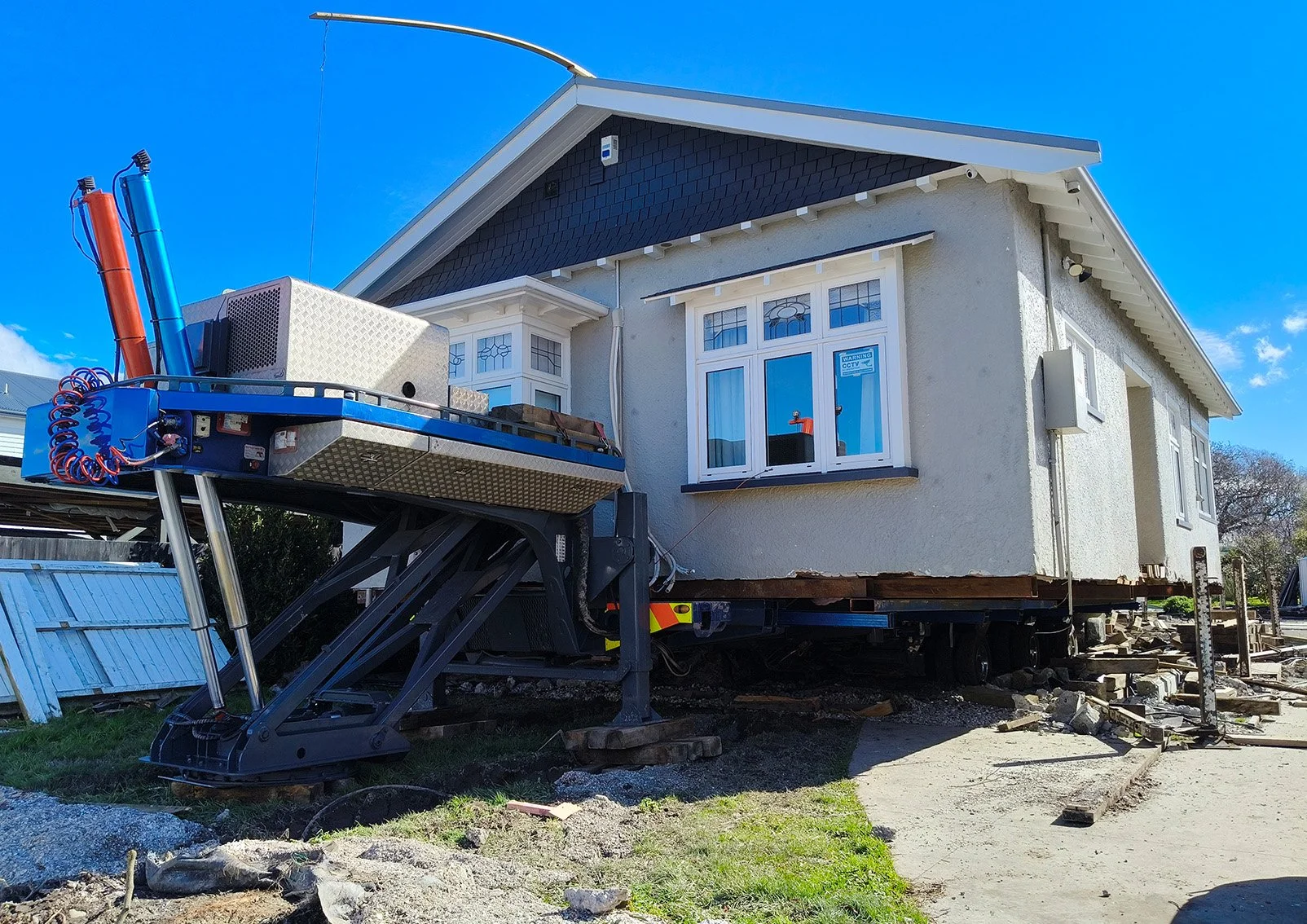A single-story character home loaded onto a heavy-duty hydraulic house moving trailer, ready for transport during a house relocation project under a clear blue sky.