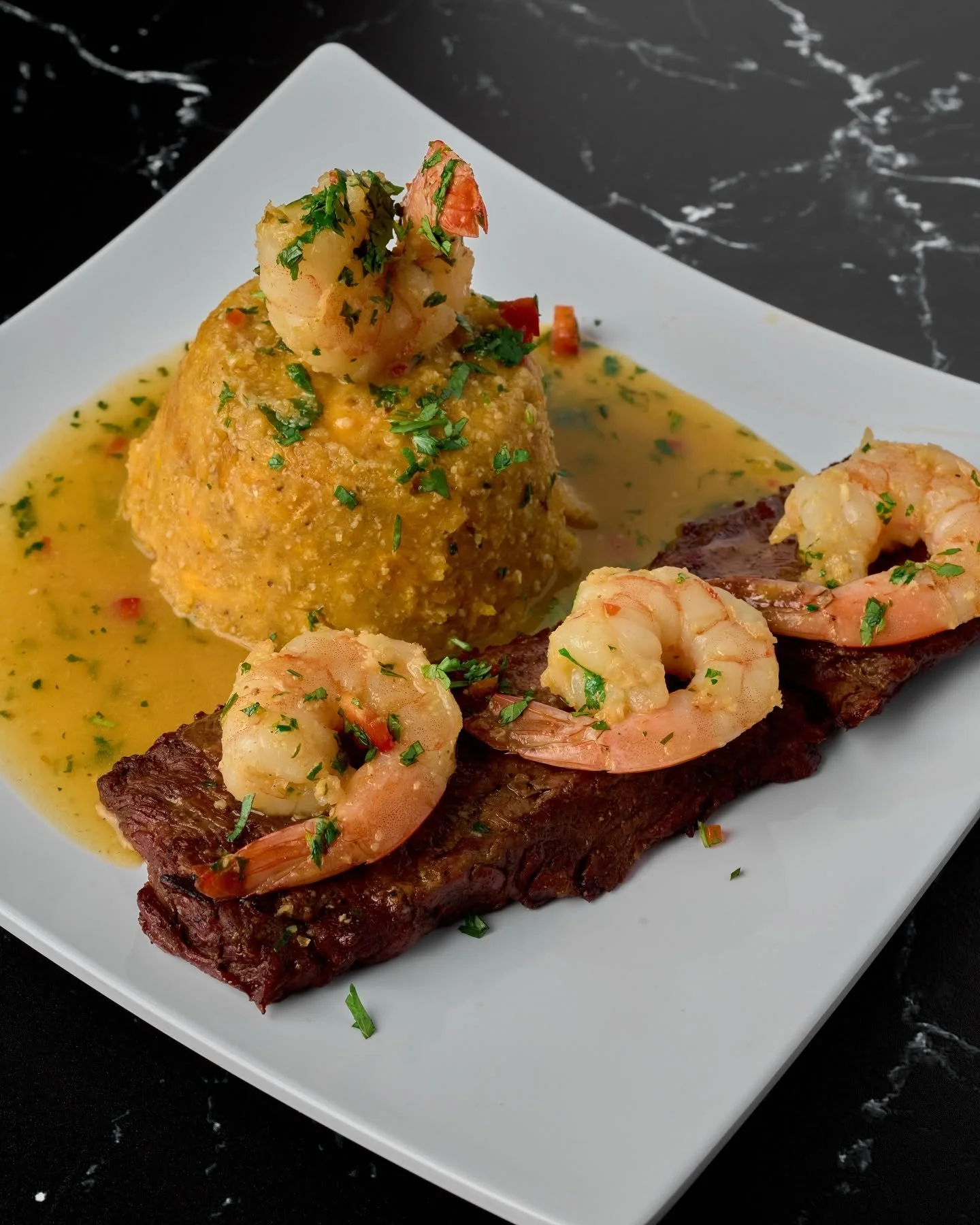 A plate of shrimp and steak topped with garlic butter and garnished with chopped parsley, with a scoop of mashed potatoes garnished with parsley.