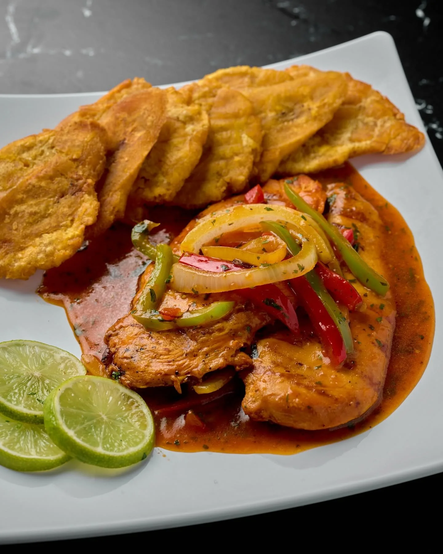 Grilled fish topped with colorful sautéed peppers, served with lime slices and fried plantains on a white plate.