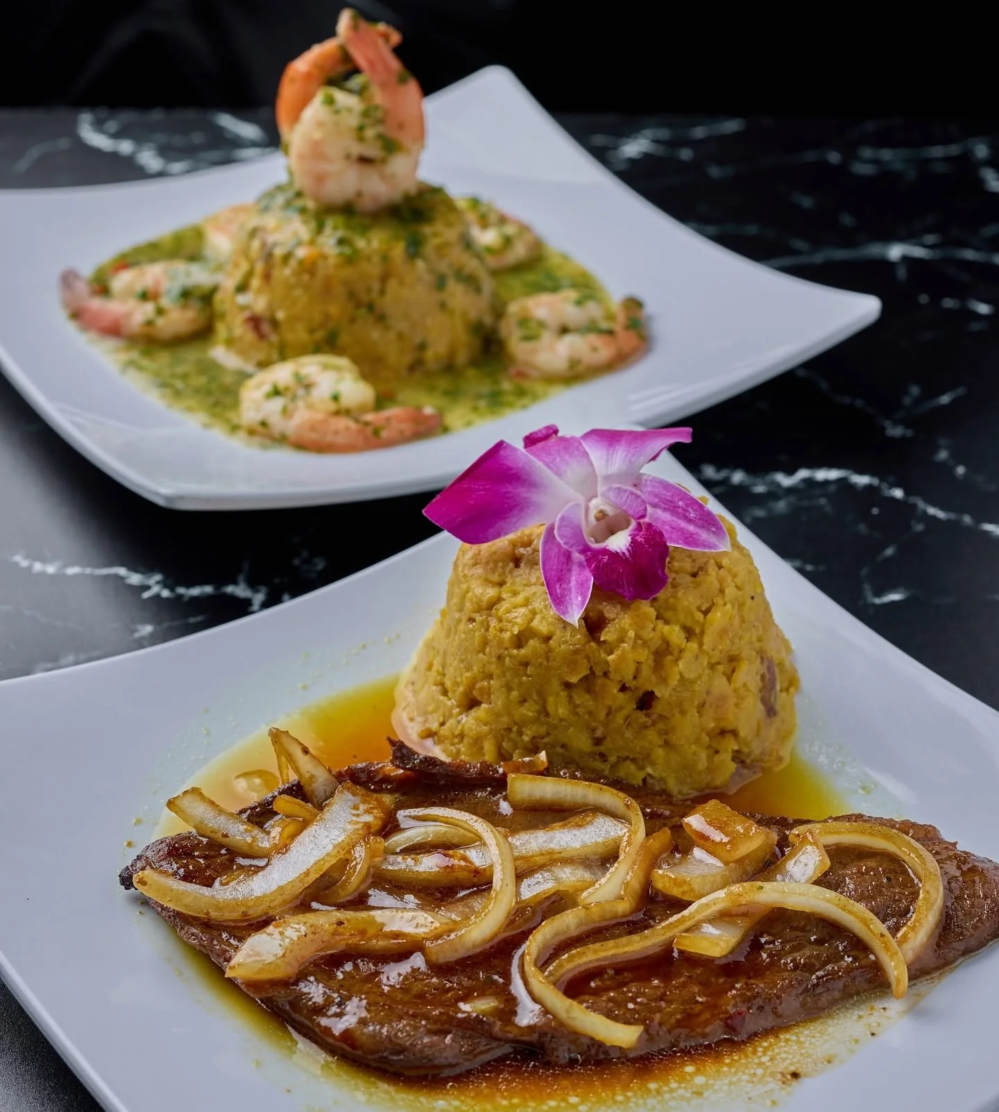 Three plated dishes of gourmet cuisine. The front plate features a breaded item topped with a pink flower, served with a savory sauce and sautéed onions. The second plate in the background has a mound of yellow rice with shrimp and a green sauce, gar