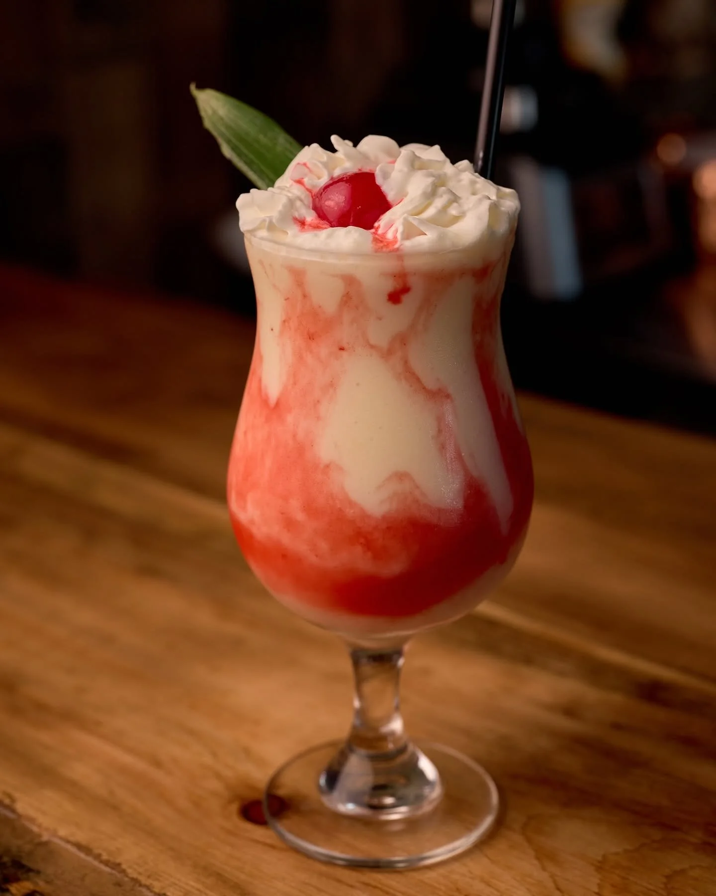 A tropical cocktail in a tall, curvy glass with whipped cream, a cherry on top, and a pineapple leaf garnish, set on a wooden table.