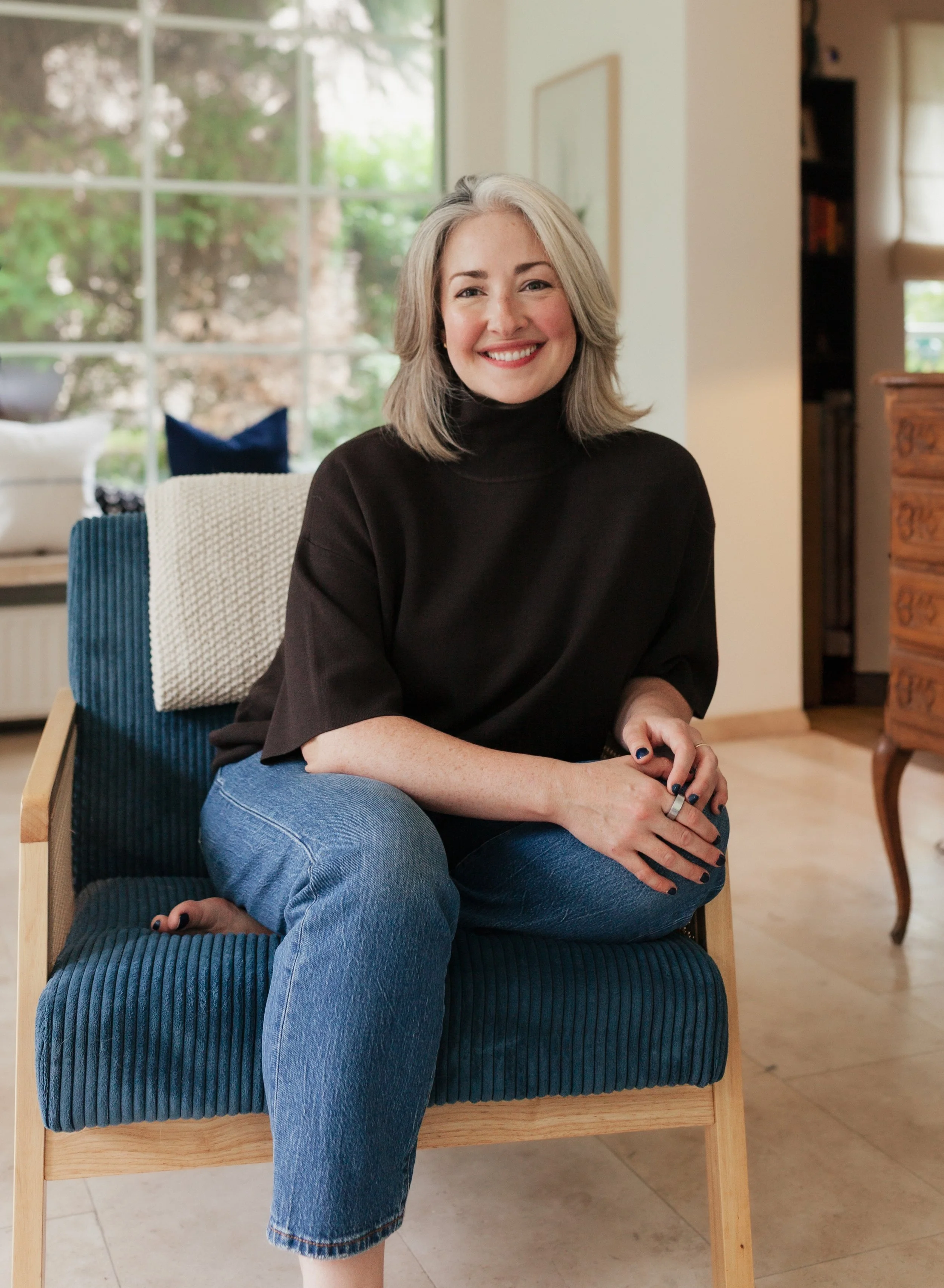Tara Berthier sitting on a wooden chair with a blue ribbed cushion in a well-lit room with large windows and a view of greenery outside.