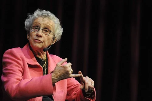 An elderly woman with curly gray hair, wearing glasses and a pink blazer, speaking into a microphone in front of a dark curtain.