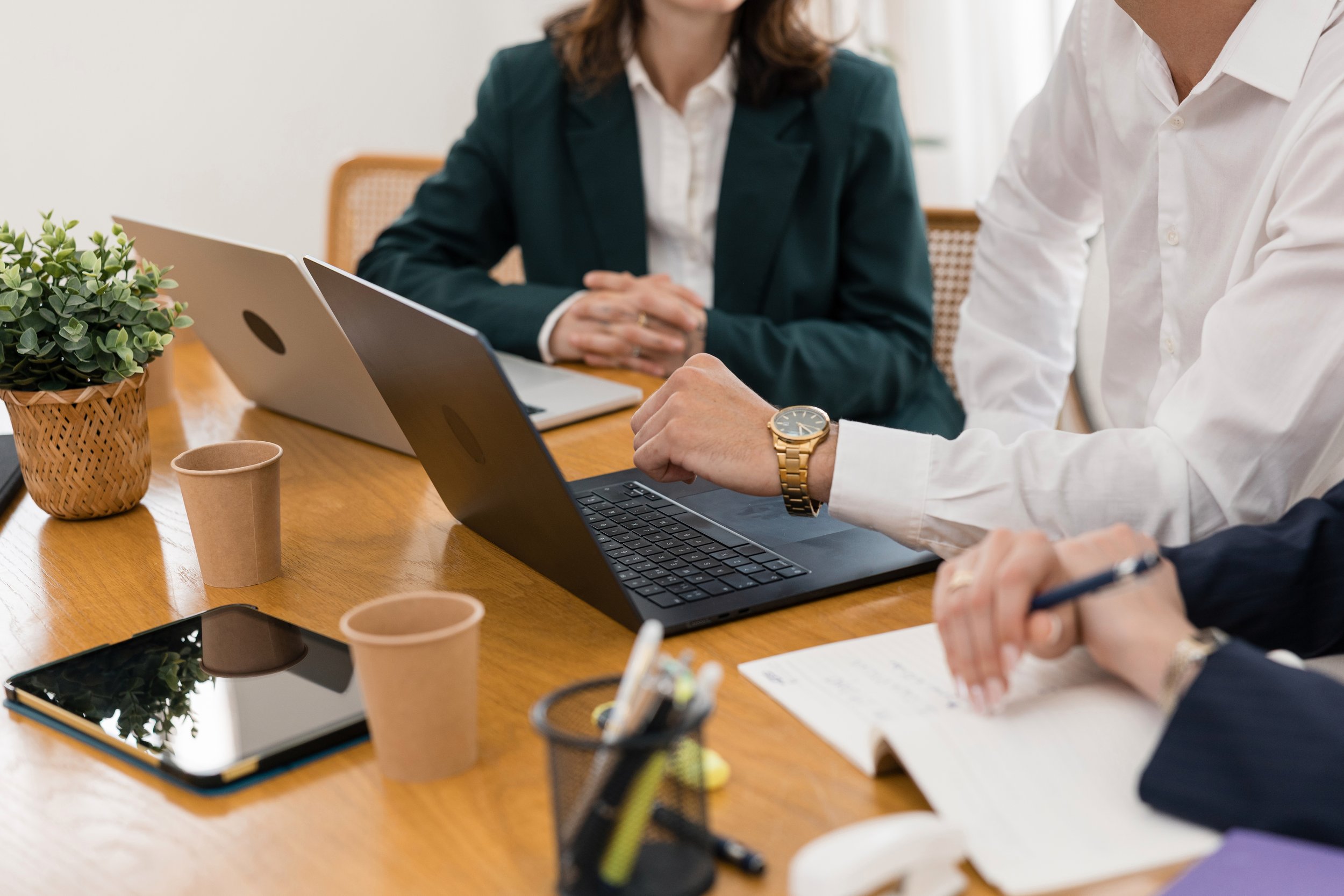 People sitting at a wooden conference table with laptops, tablets, and papers, during a meeting in a bright room.