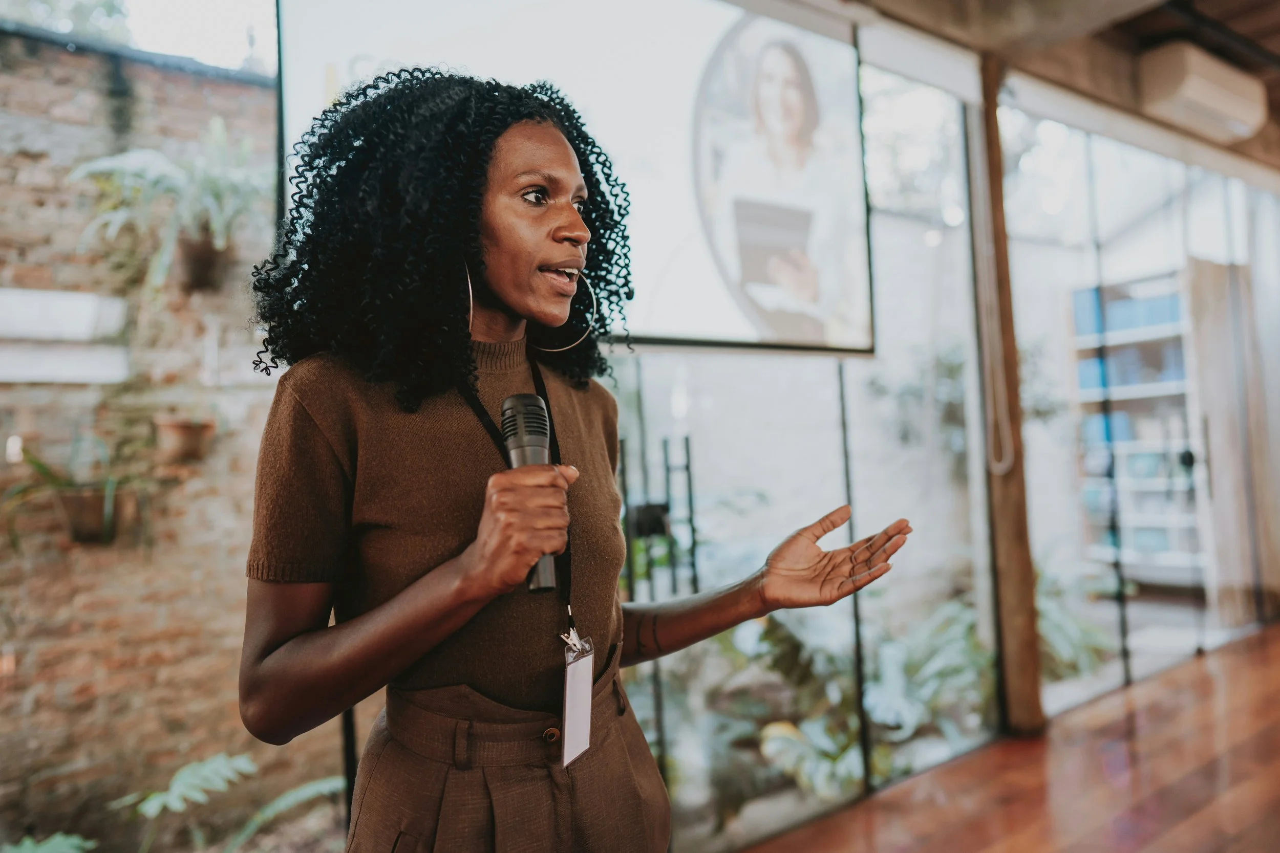 A woman with curly black hair, wearing a brown top, speaking into a microphone in what appears to be a conference or seminar with a brick wall and large windows in the background.