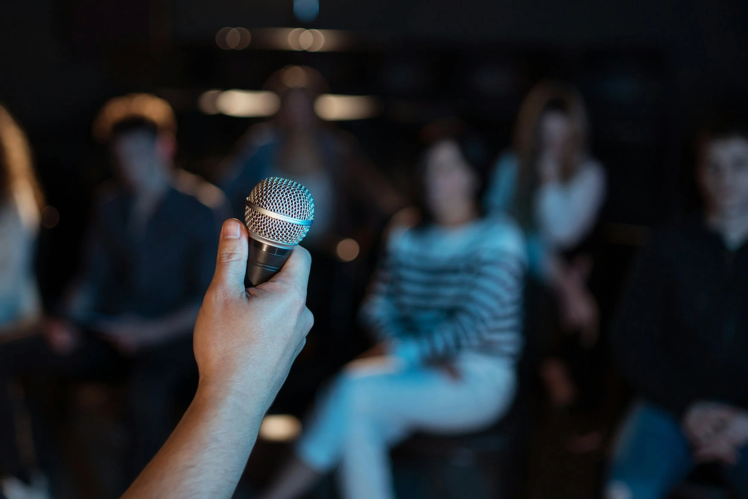 Person holding a microphone in front of an audience of people sitting in a dimly lit room.