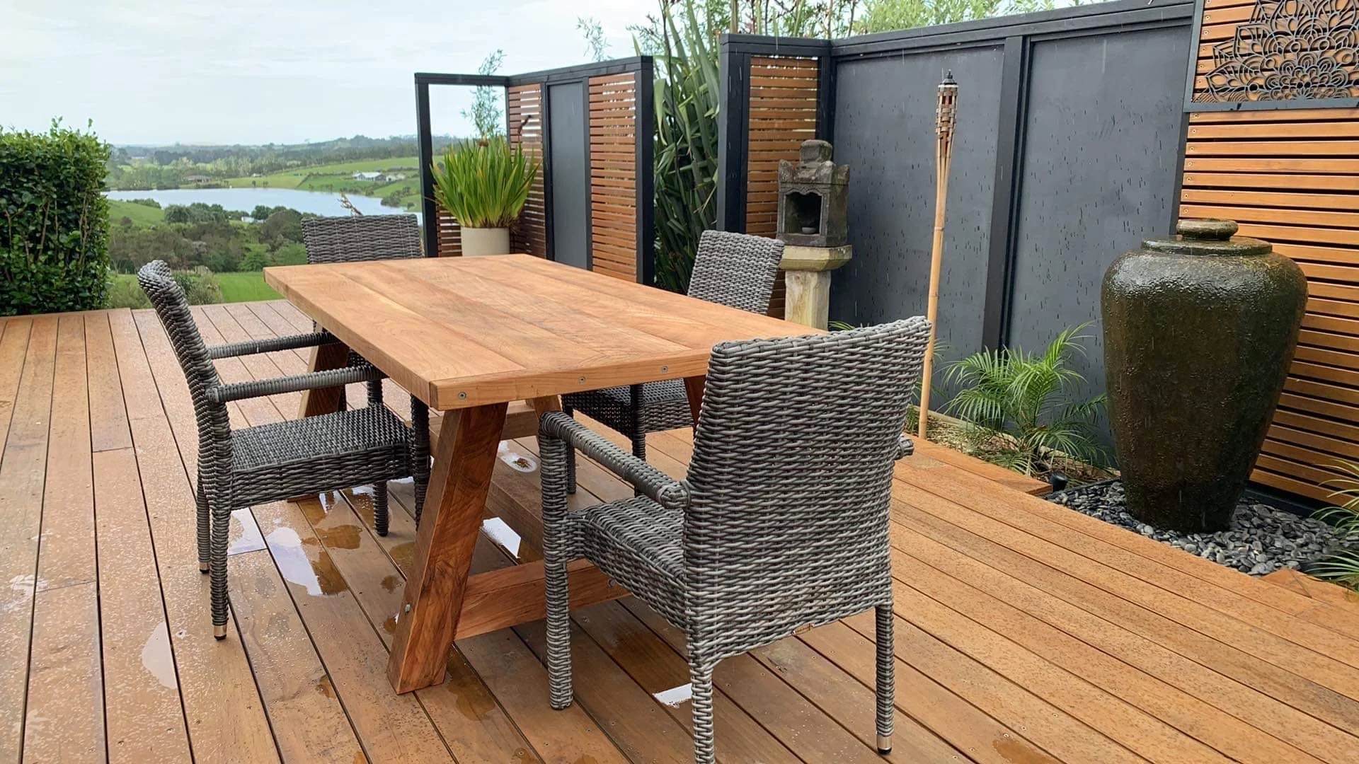Macrocarpa outdoor dining table with woven chairs on a deck overlooking the water.