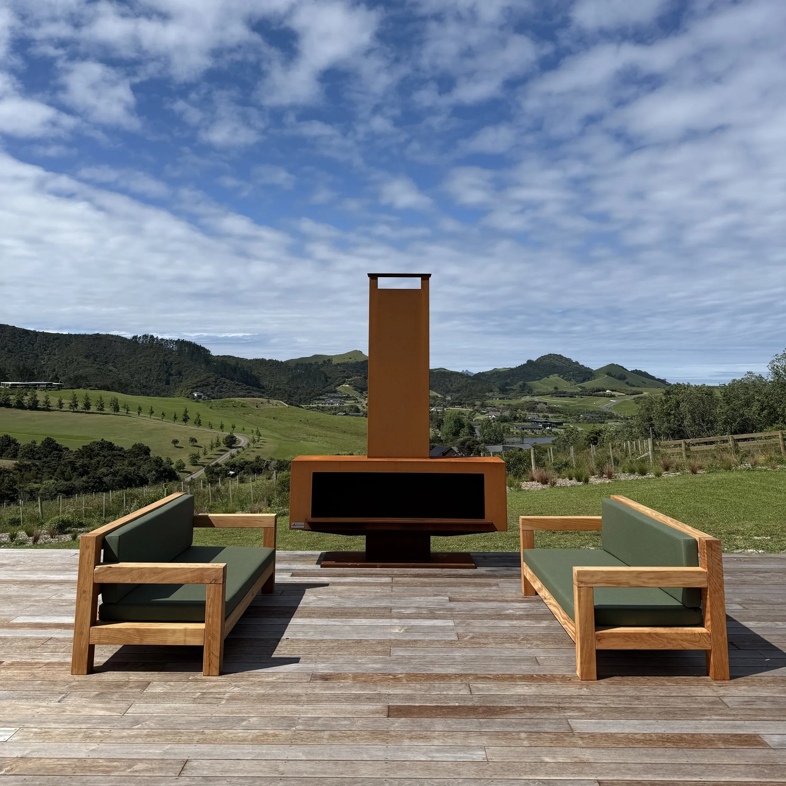 Two Macrocarpa outdoor lounge seats with green squabs beside an outdoor fireplace.