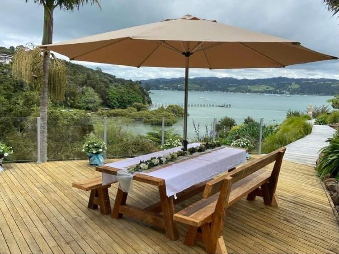Macrocarpa dining table and matching benches beneath an outdoor umbrella overlooking the water.