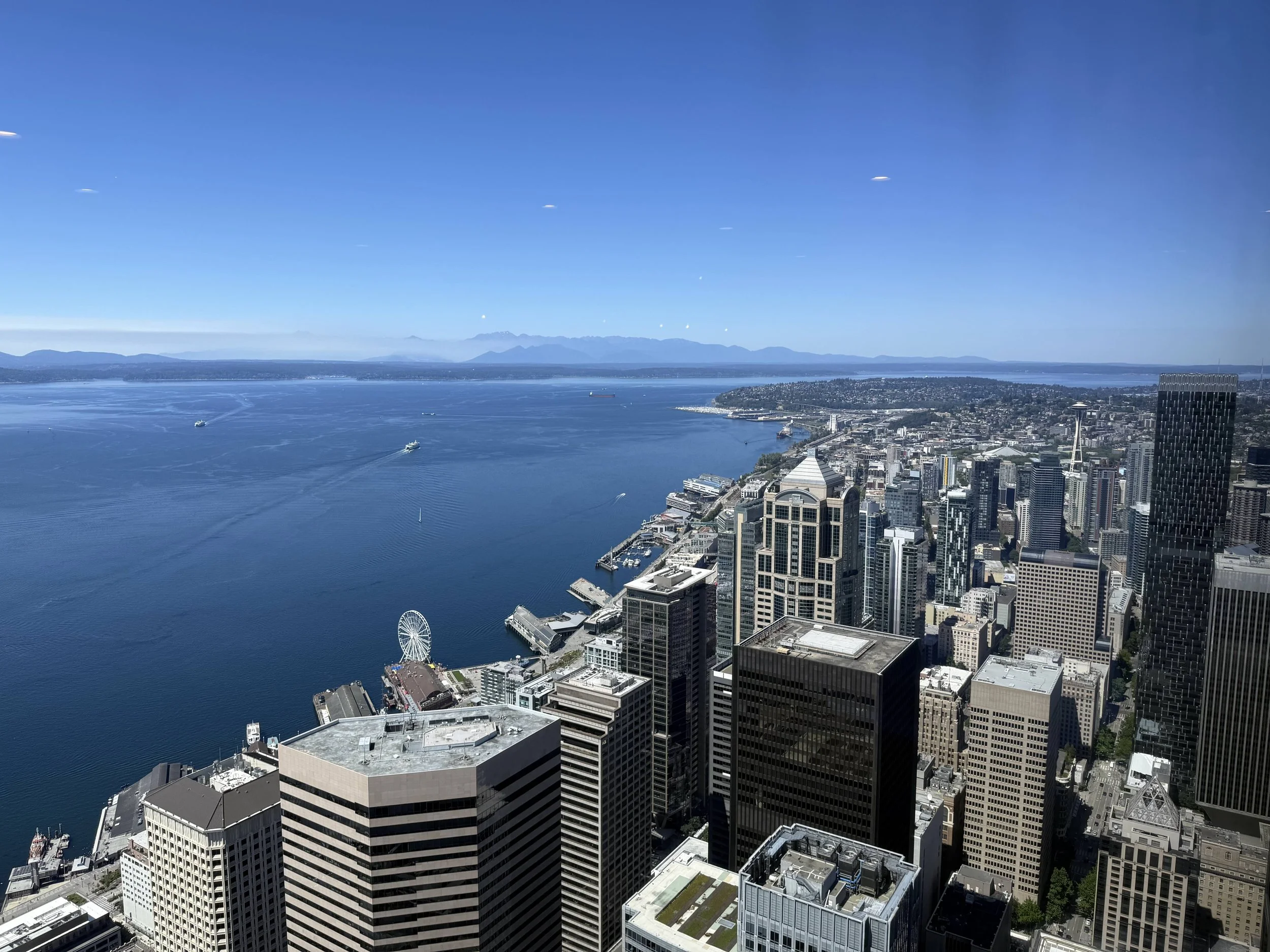 A high-angle cityscape view of Seattle, Washington, featuring tall skyscrapers, Elliott Bay, boats, and the Seattle Space Needle in the distance under a clear blue sky.