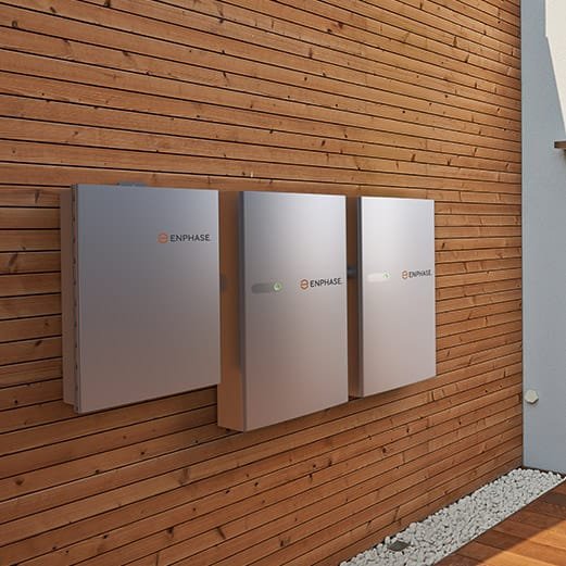 Three gray enphase solar battery storage units mounted on a wooden wall outside.