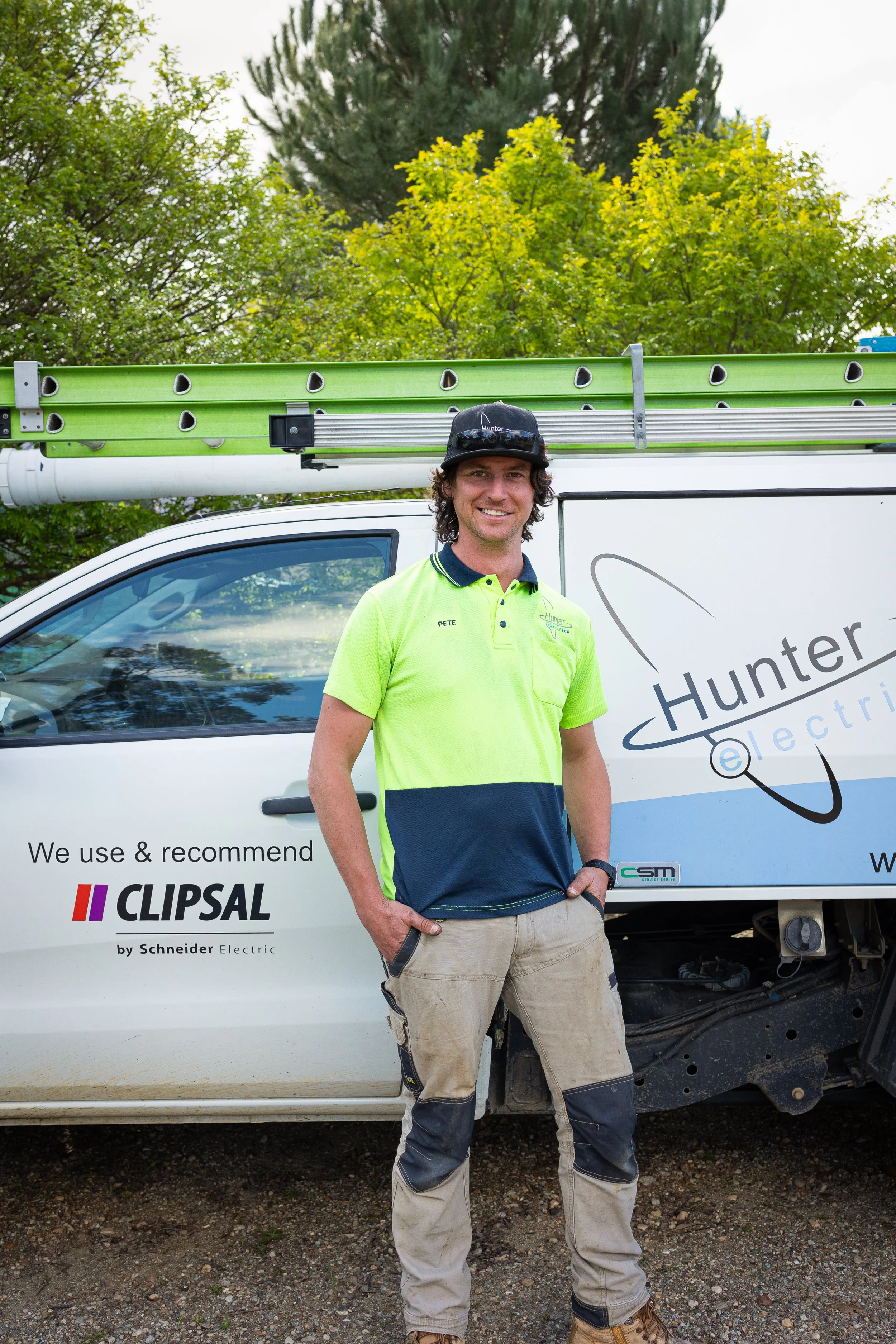Man standing in front of a service truck with ladder, wearing a high visibility yellow and navy shirt, khaki work pants, and sunglasses on his head, outdoors with trees in the background.