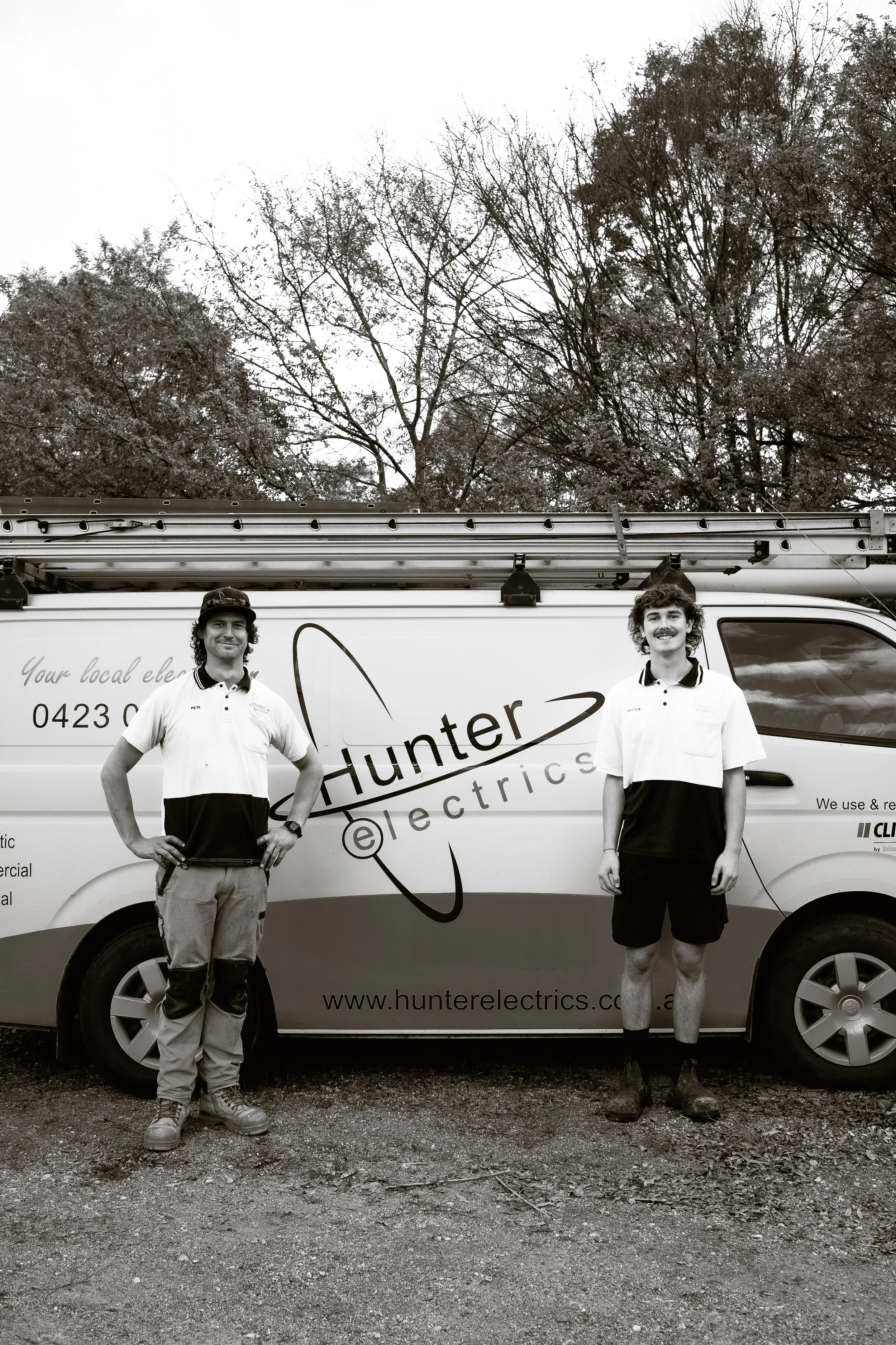 Two young men standing in front of a van with company branding for Hunter Electrics, holding work tools, outdoors with trees in the background.