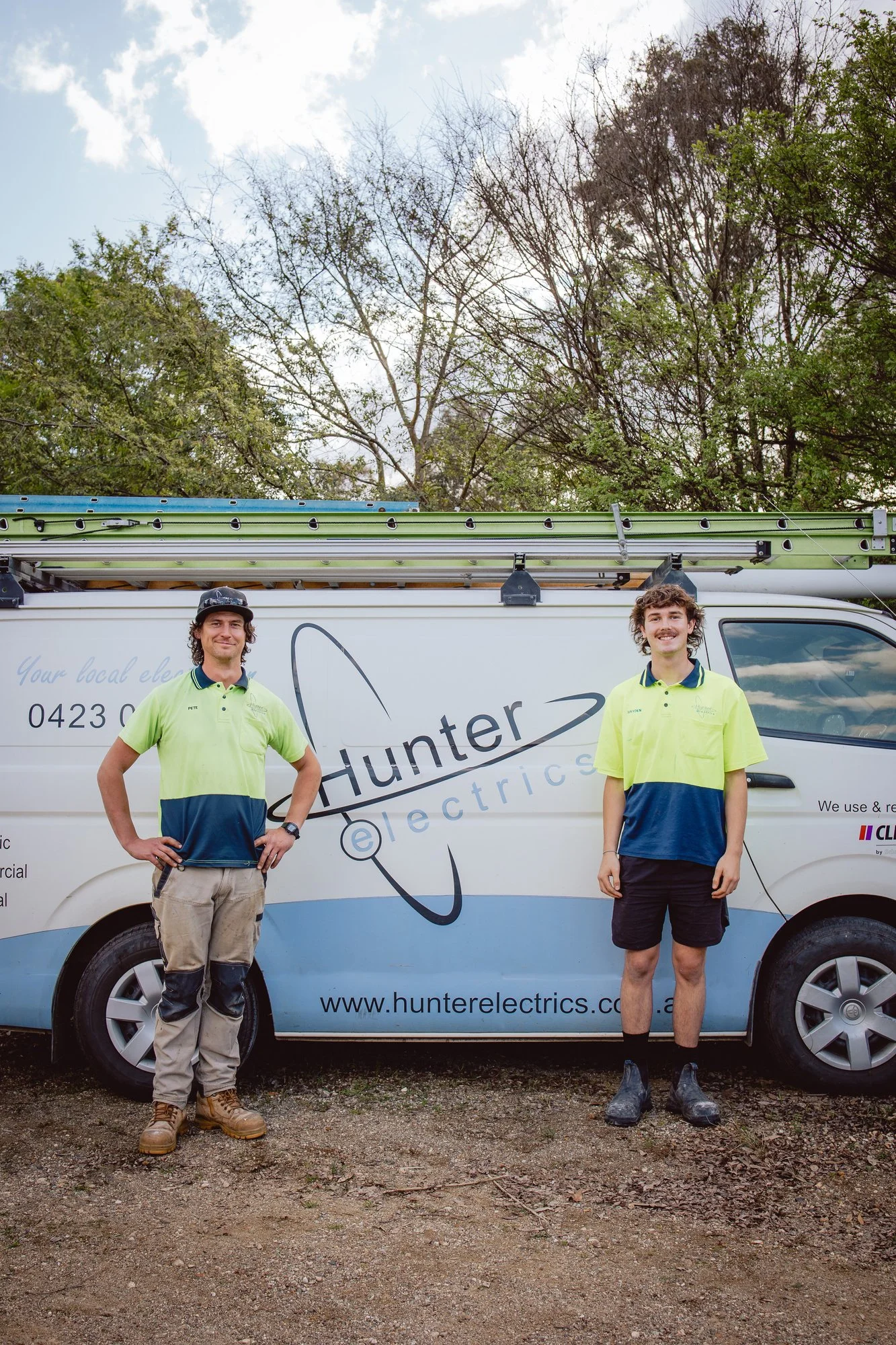 Two young men standing in front of a company van with the logo 'Hunter Electrics.' The man on the left is wearing work gear, and the man on the right is dressed in safety clothing. The background includes trees and a partly cloudy sky.
