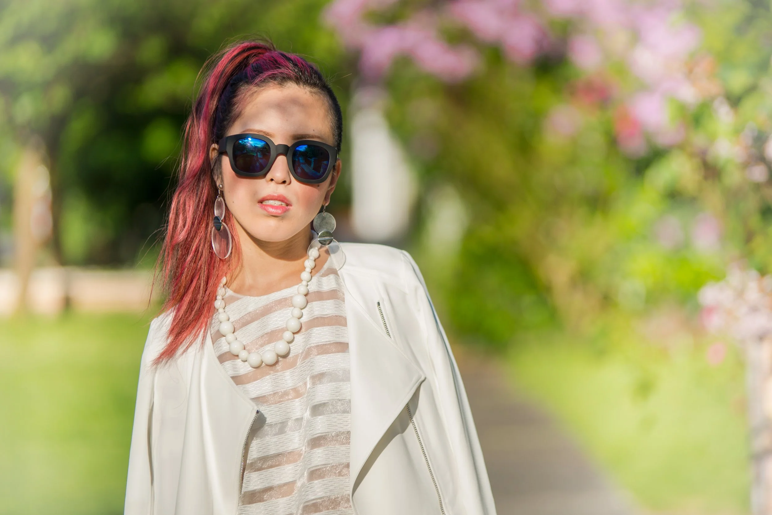A woman with pink streaked hair wearing black sunglasses, large earrings, a white beaded necklace, and a white jacket, standing outdoors with blurred green and pink foliage in the background.